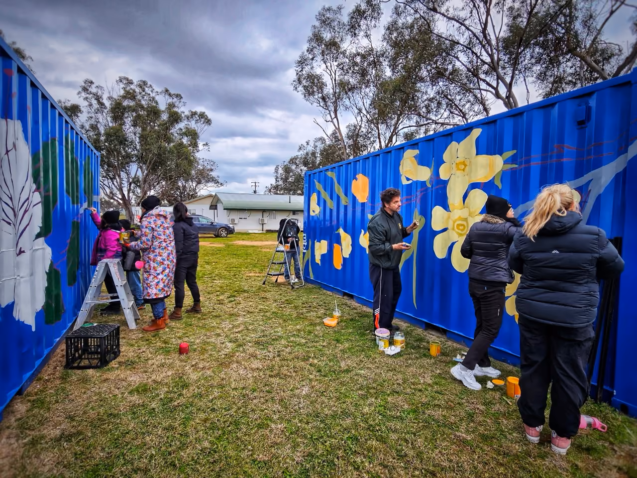 Six people painting colorful floral murals on blue shipping containers outdoors on a cloudy day.