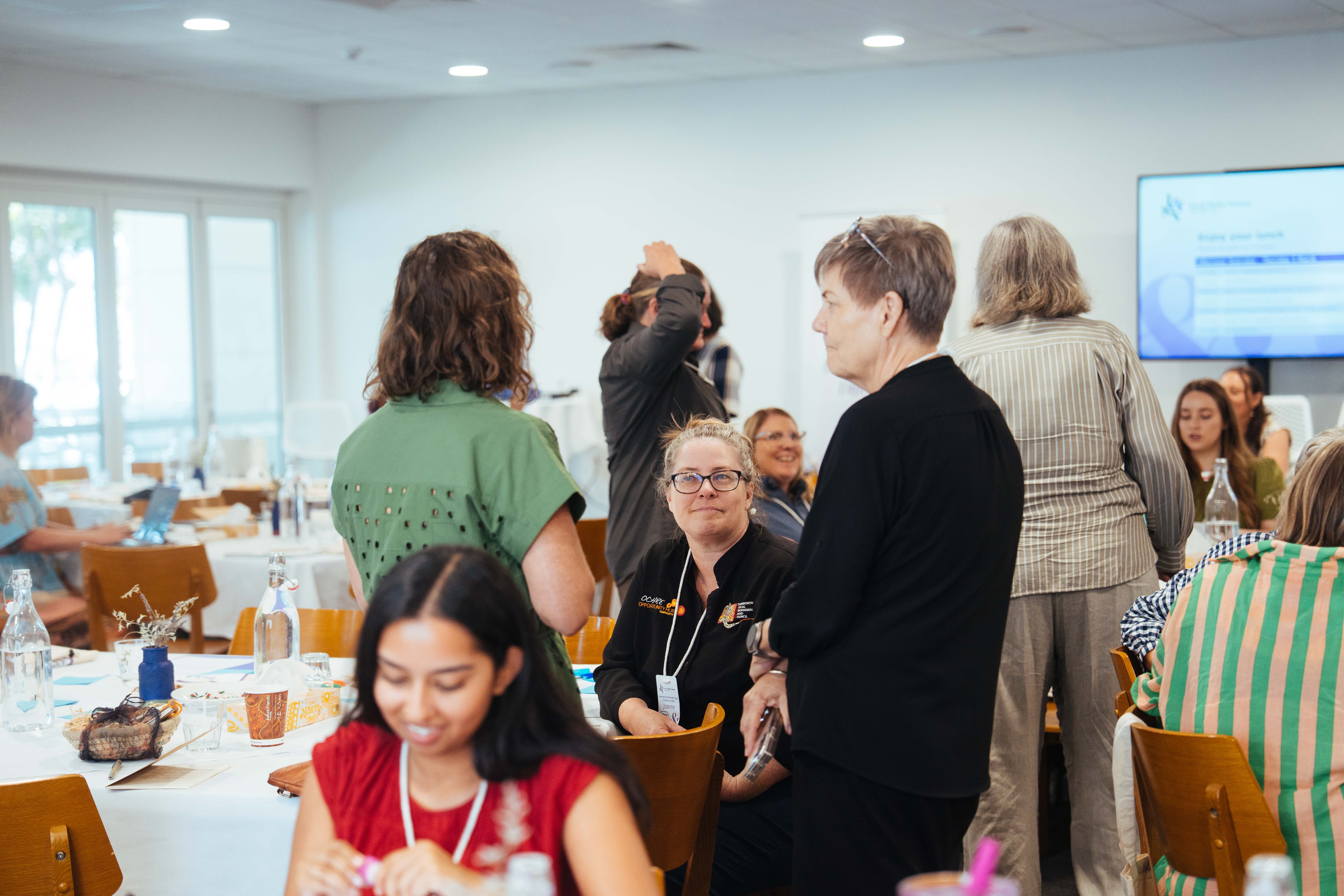 Group of diverse women talking and sitting around tables in a bright meeting room.