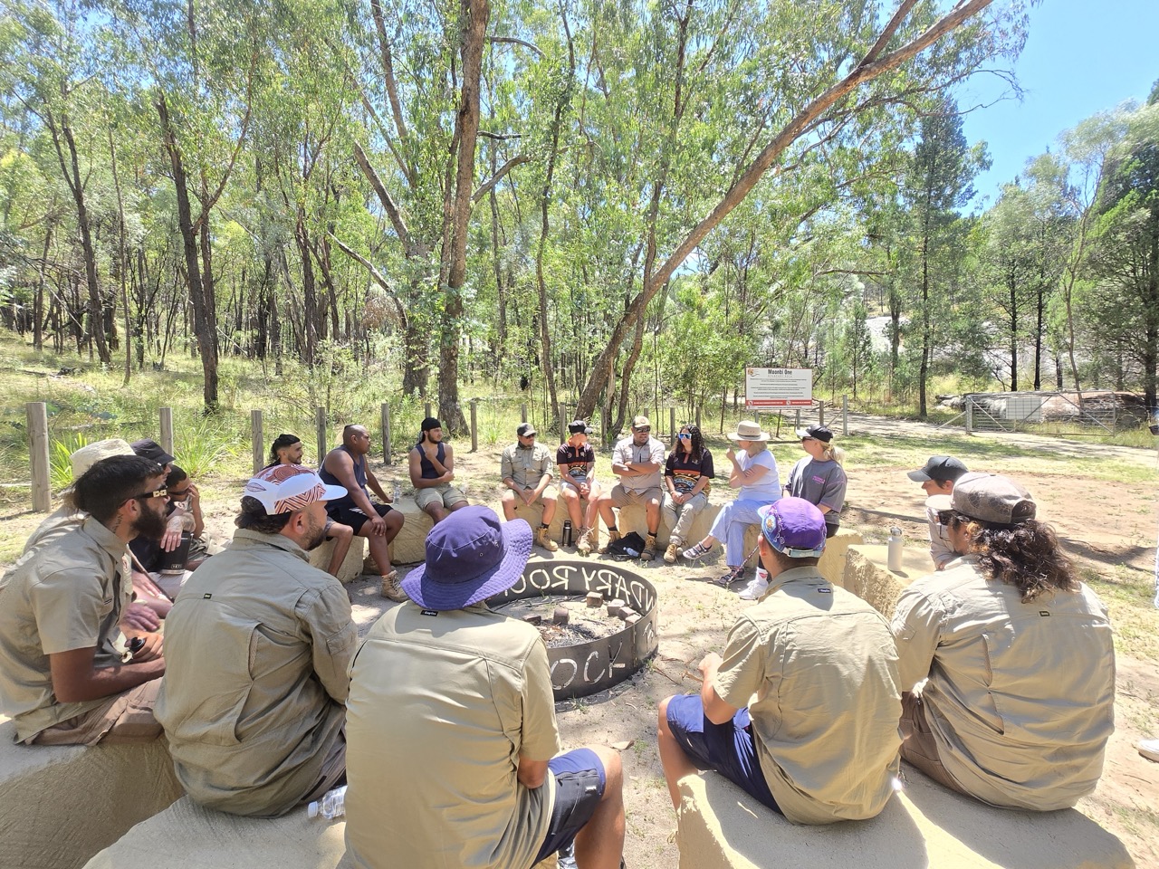 Group of people sitting in a circle outdoors around a fire pit in a wooded area on a sunny day.