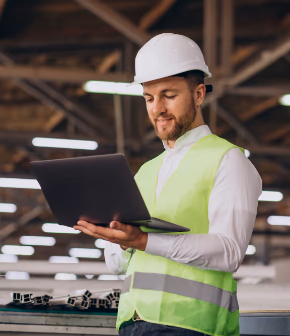Man wearing white hard hat and yellow safety vest using laptop in industrial warehouse.