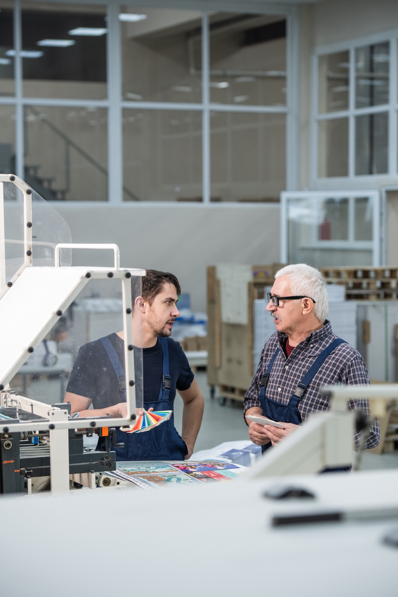 Two male workers in overalls discussing prints and color samples in a printing factory.