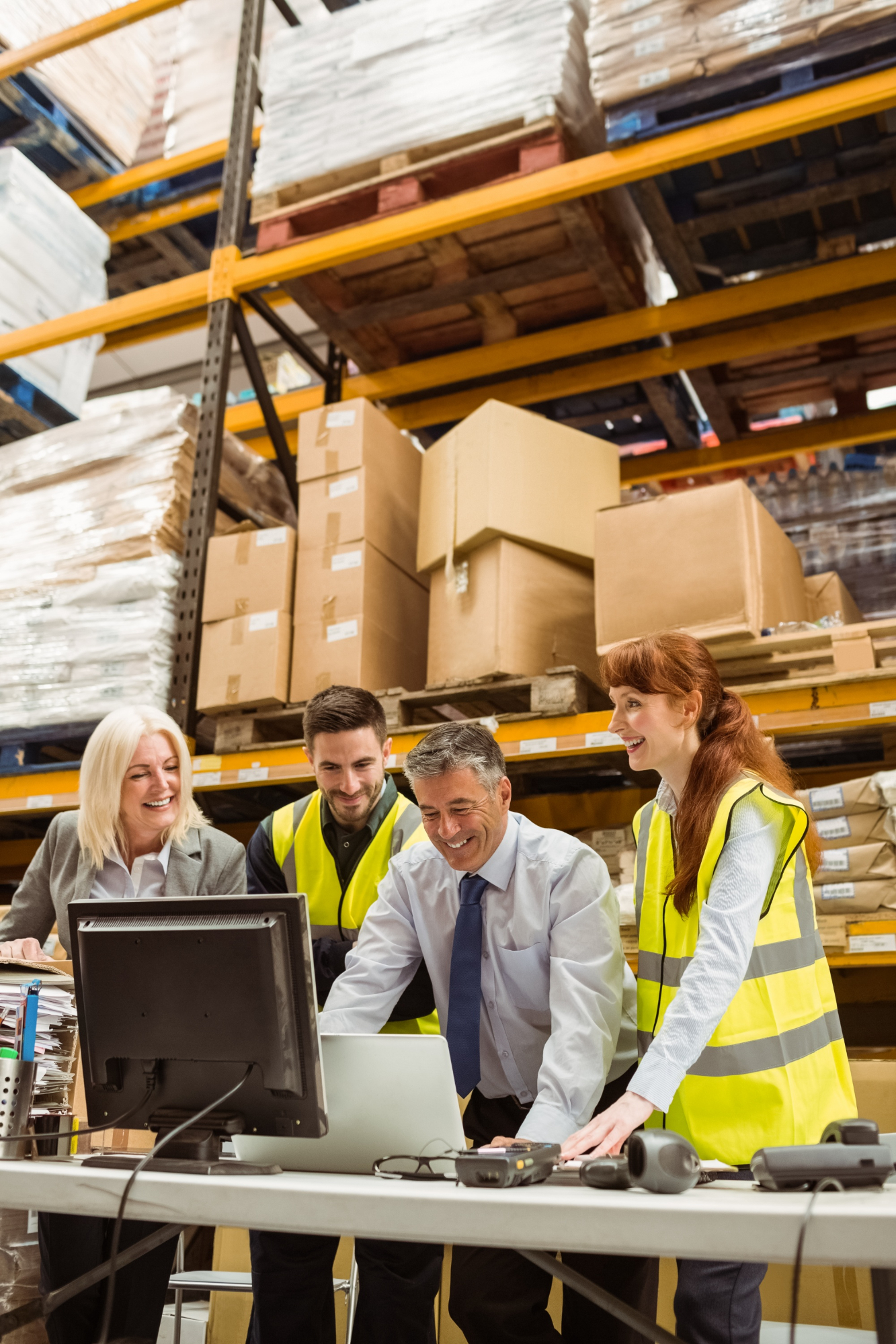 Four warehouse workers smiling and collaborating at a desk with computer monitors and packages.