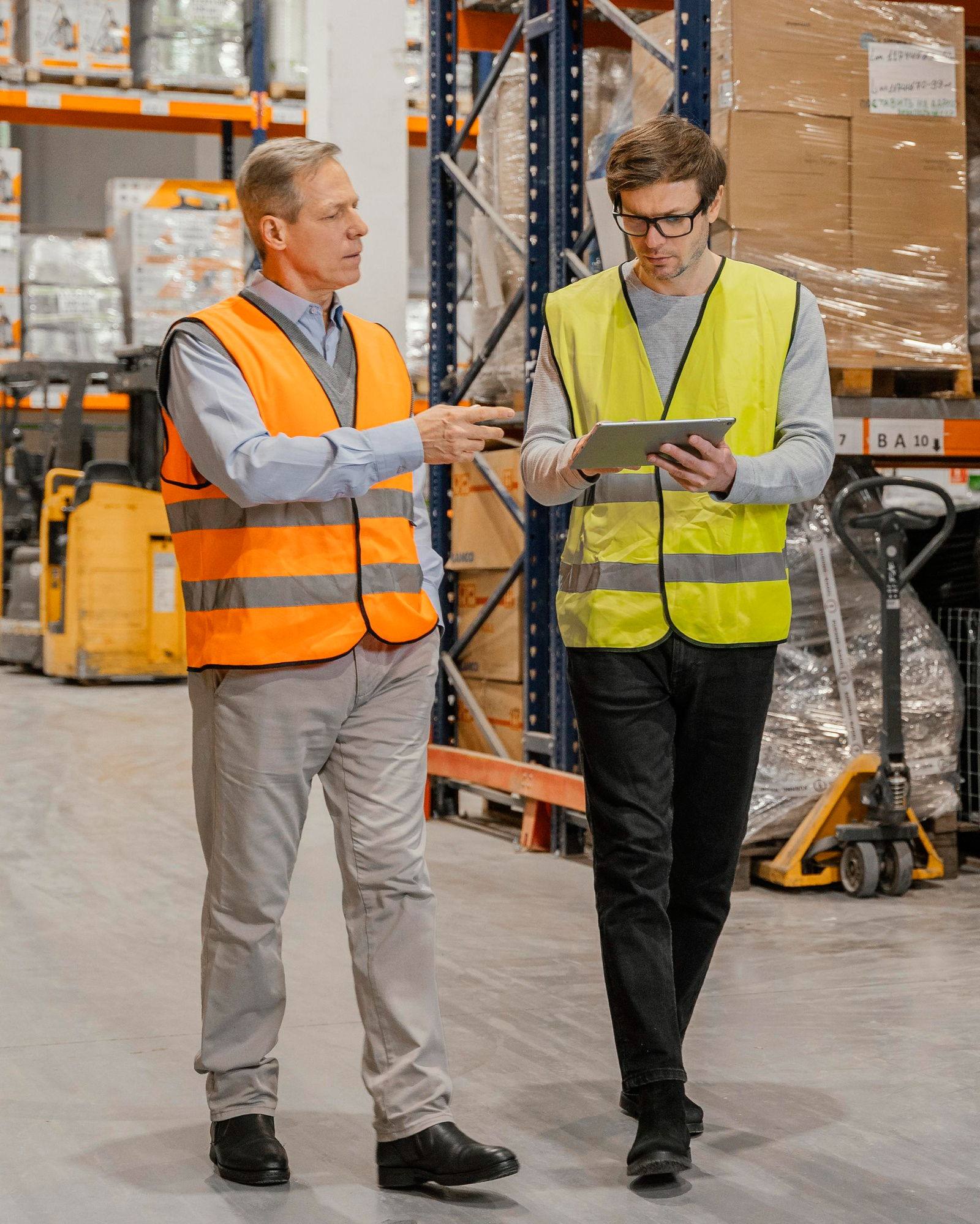 Two warehouse workers in safety vests discussing while using a tablet in a storage aisle.