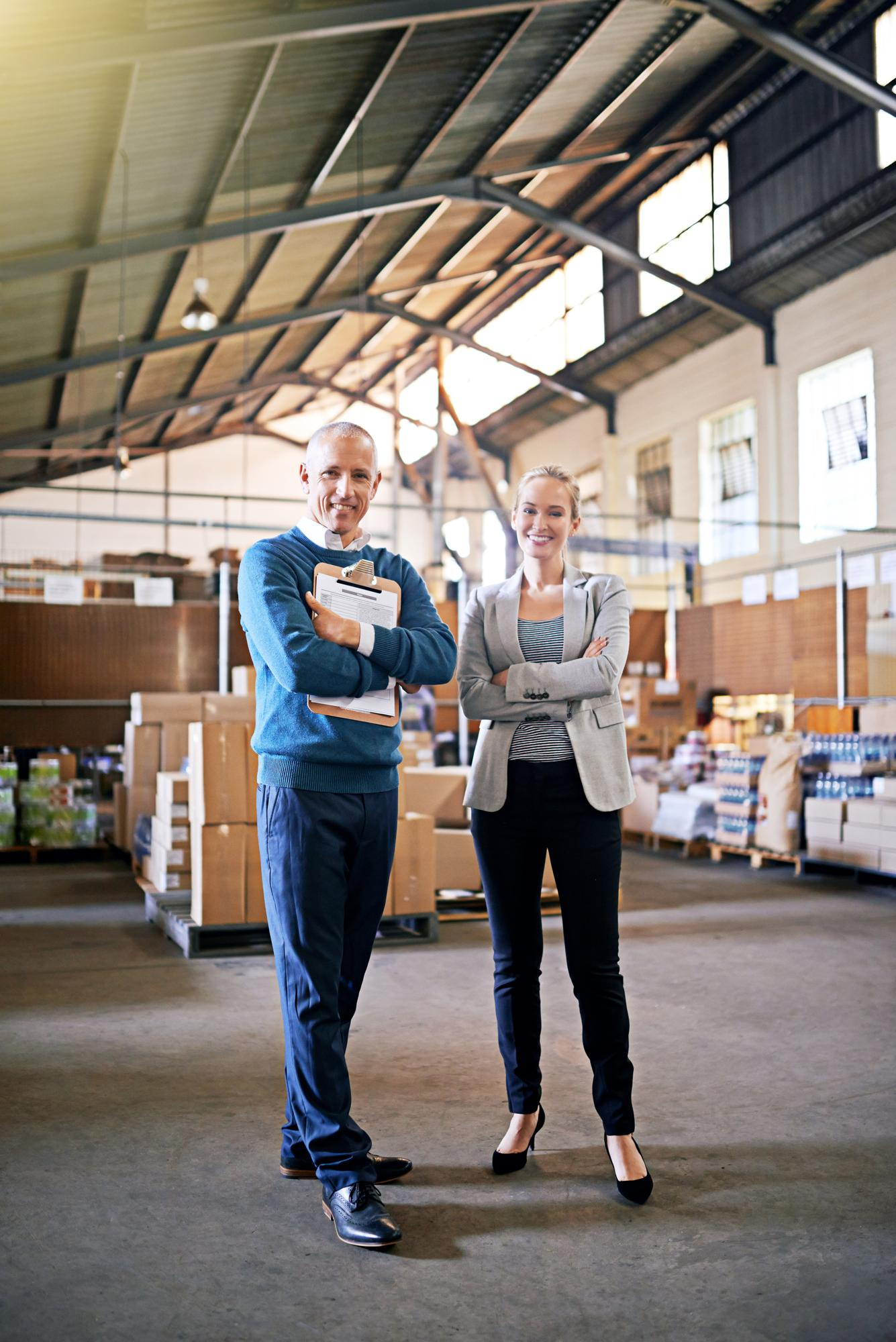 Two business professionals standing with arms crossed in a spacious warehouse with stacked boxes.