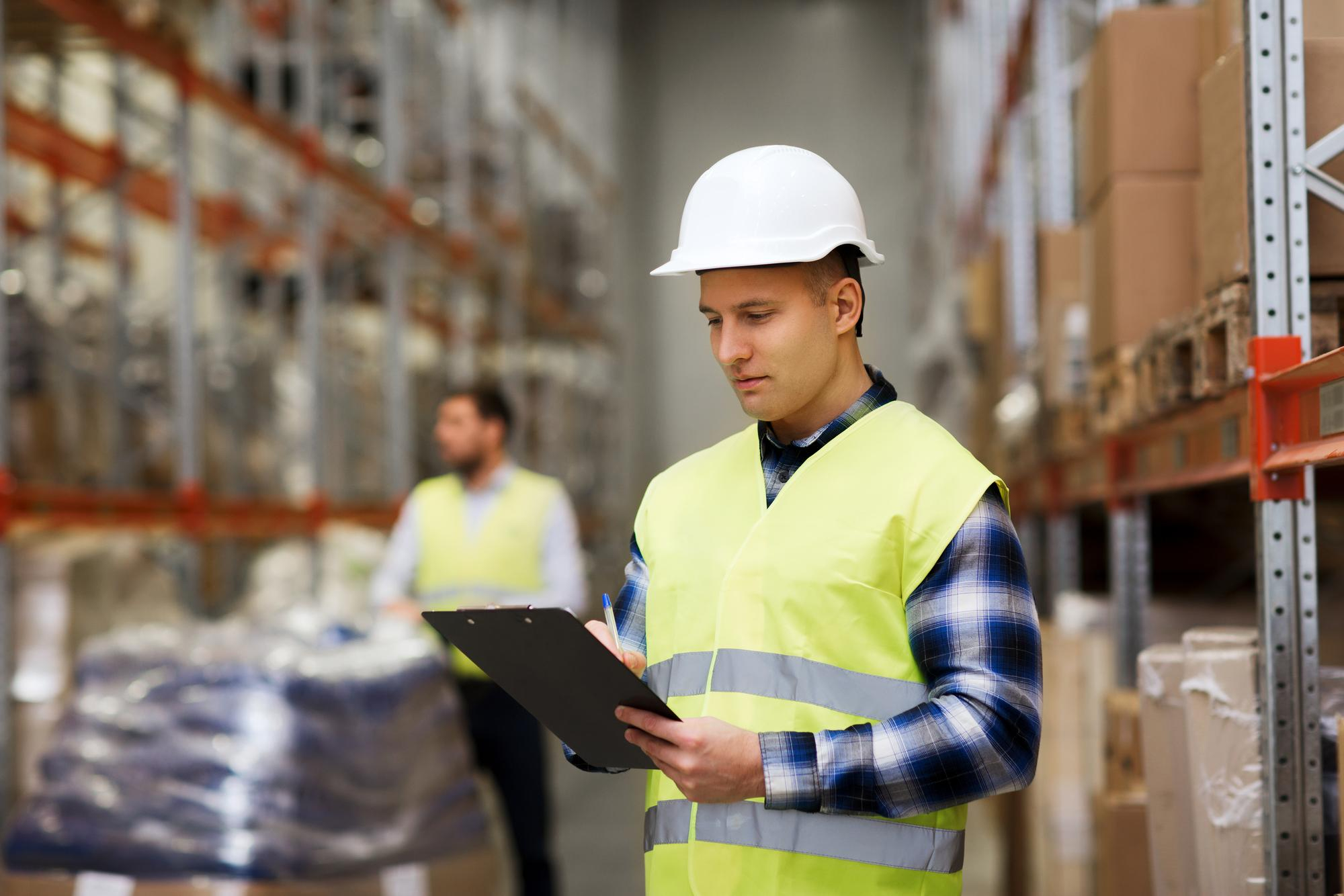 Warehouse worker in a hard hat and safety vest checks inventory on a clipboard among shelves.