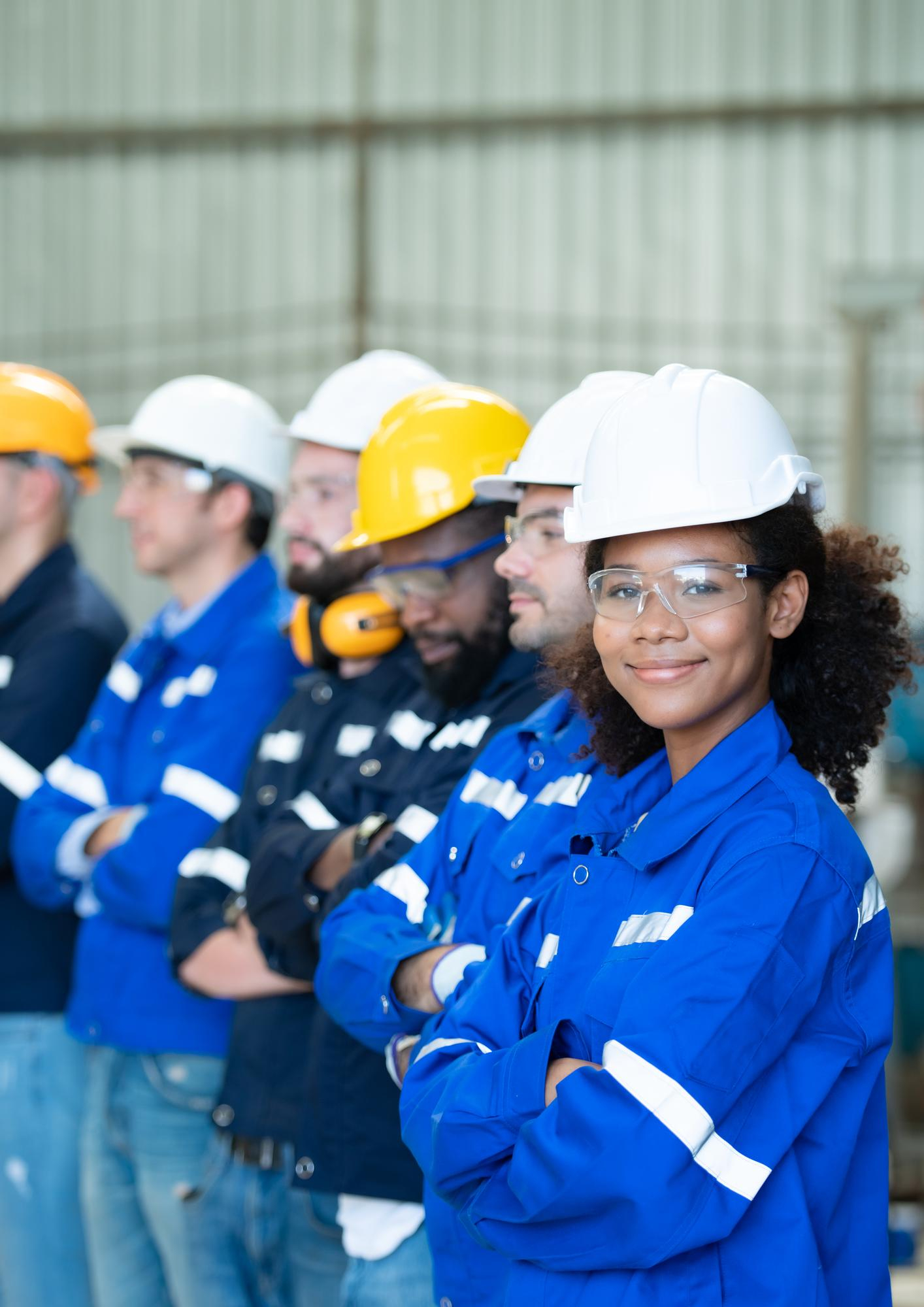 Portrait of diverse industrial workers in hard hats and blue uniforms standing with arms crossed.