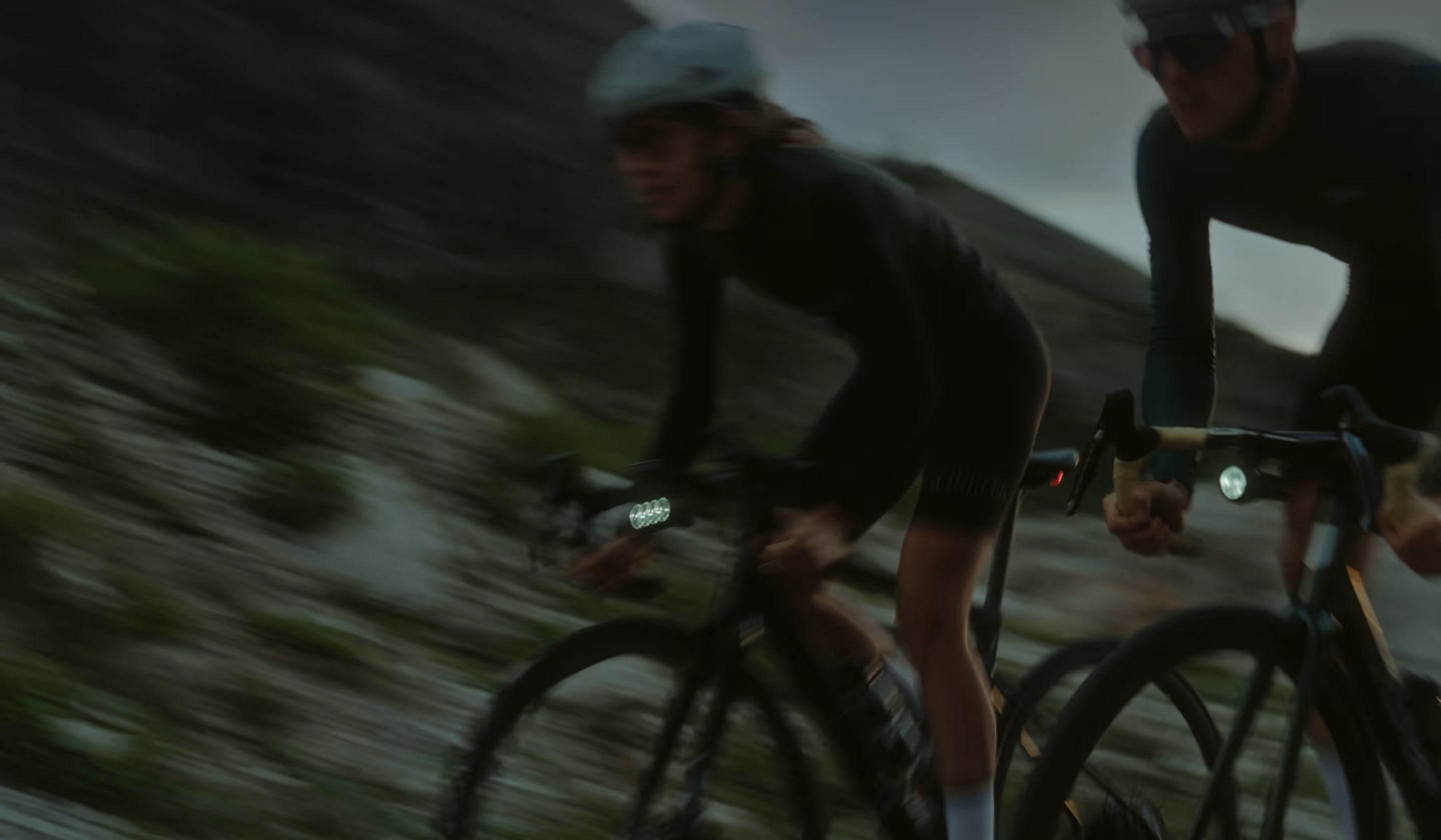 Two cyclists in helmets riding mountain bikes uphill on a rocky trail at dusk.
