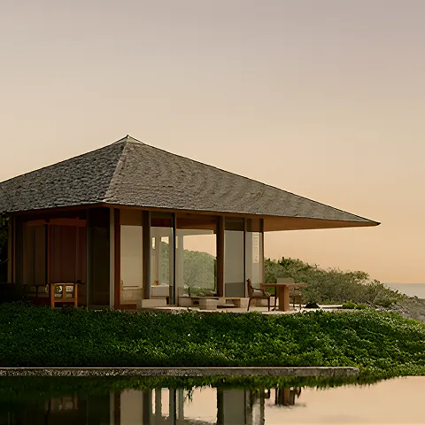 Modern pavilion with slanted roof surrounded by greenery and reflected in calm water at sunset.