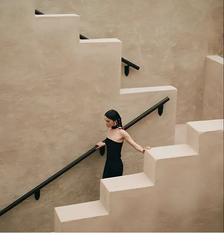 Woman in a black dress descending beige stairs with black handrails against a textured wall.