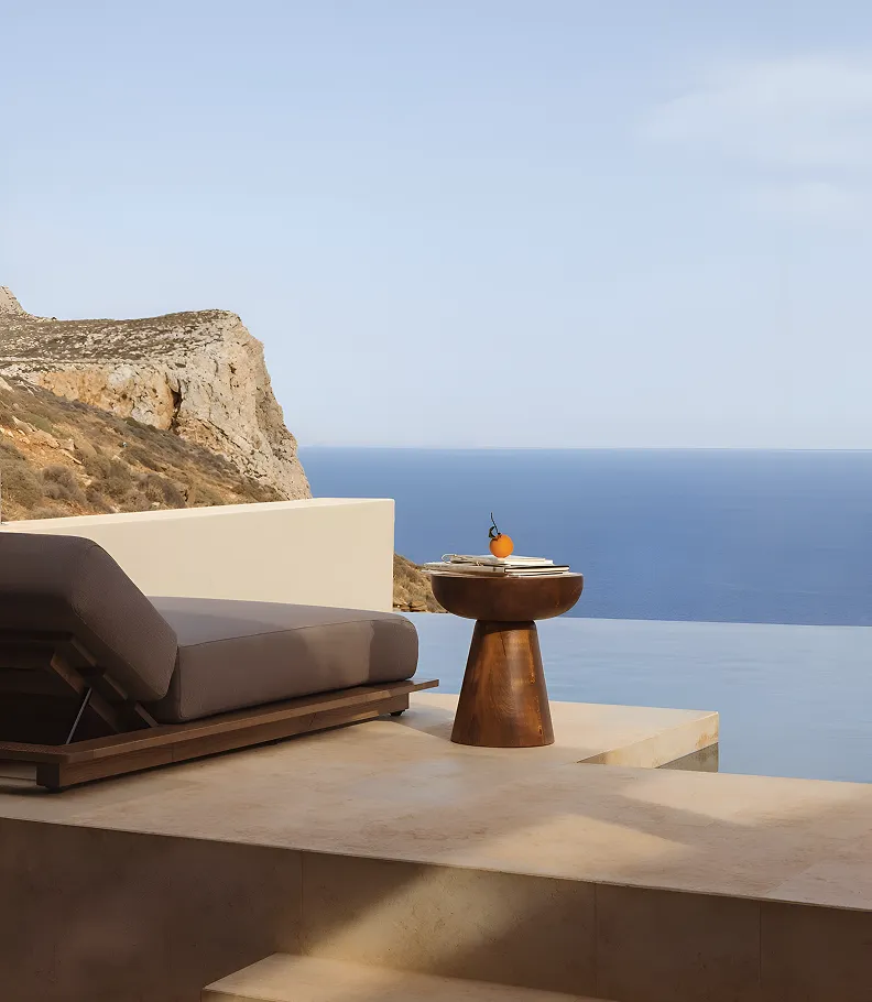 Modern lounge chair and wooden side table on terrace overlooking blue sea and rocky cliffs.
