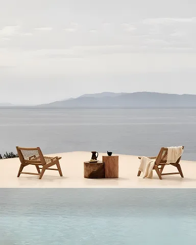 Two wooden chairs with a blanket beside a pool overlooking calm water and distant hills under a cloudy sky.
