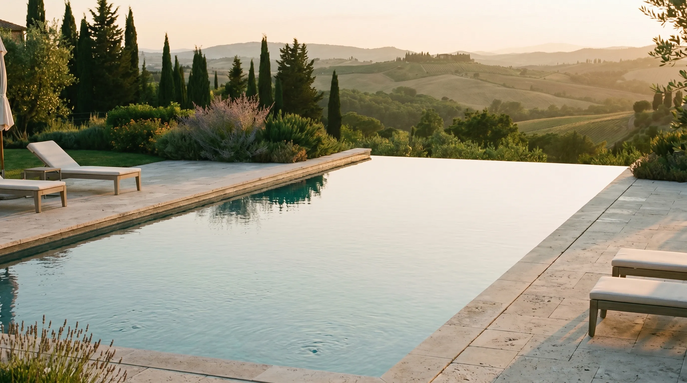 Infinity pool with lounge chairs overlooking rolling hills and trees at sunset.