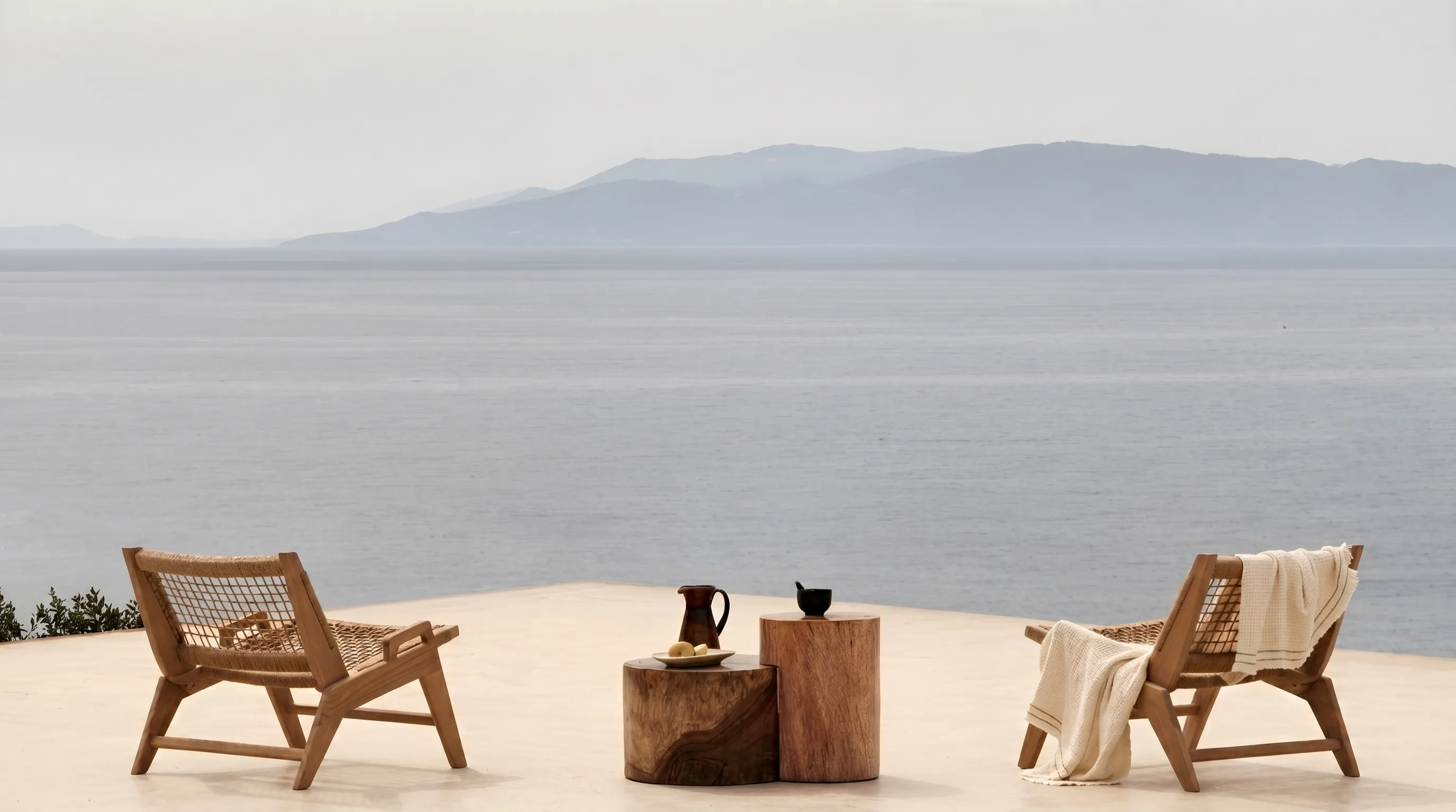 Two wooden lounge chairs and a side table on a terrace overlooking calm sea and distant mountains.