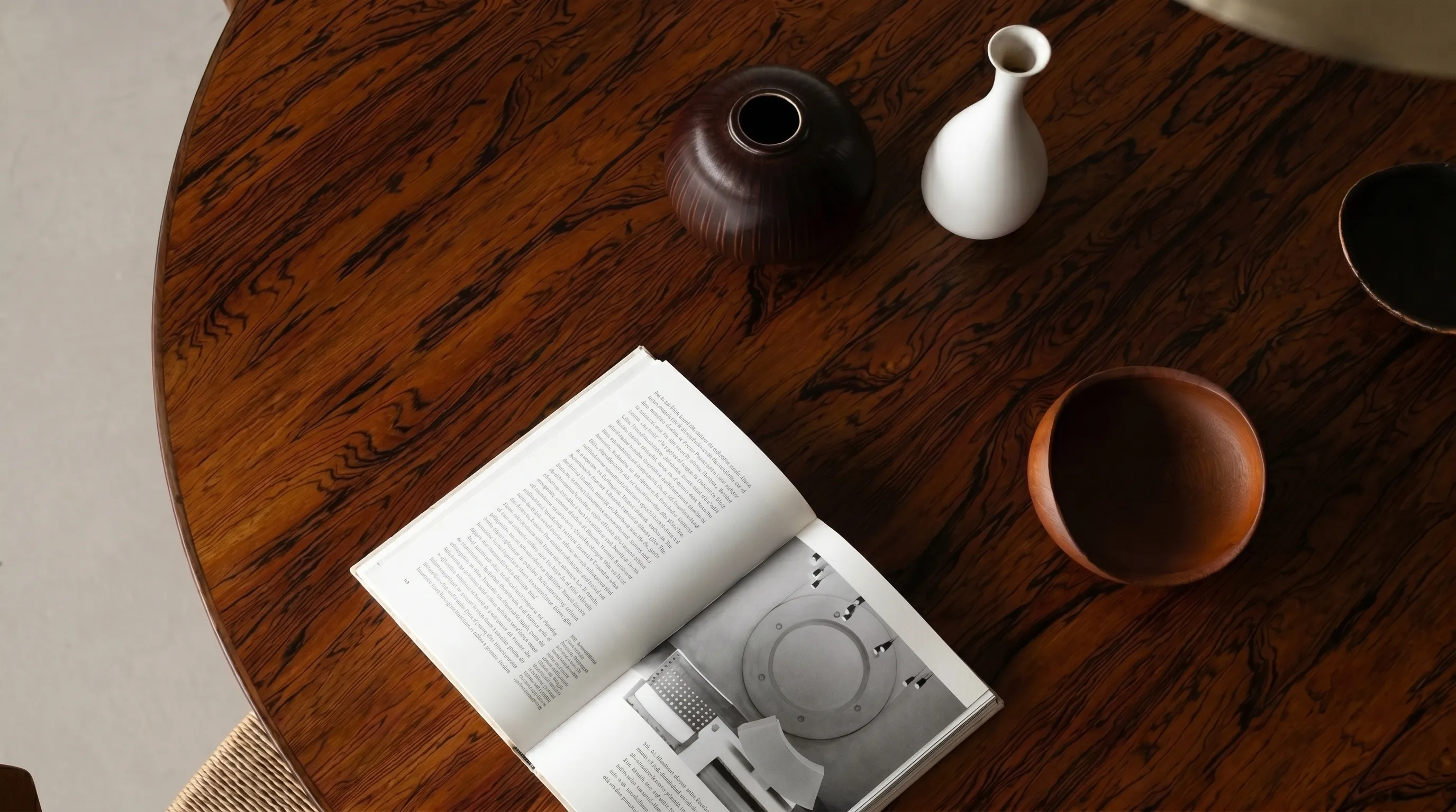 Open book with text and photograph on wooden table next to ceramic vases and wooden bowl.