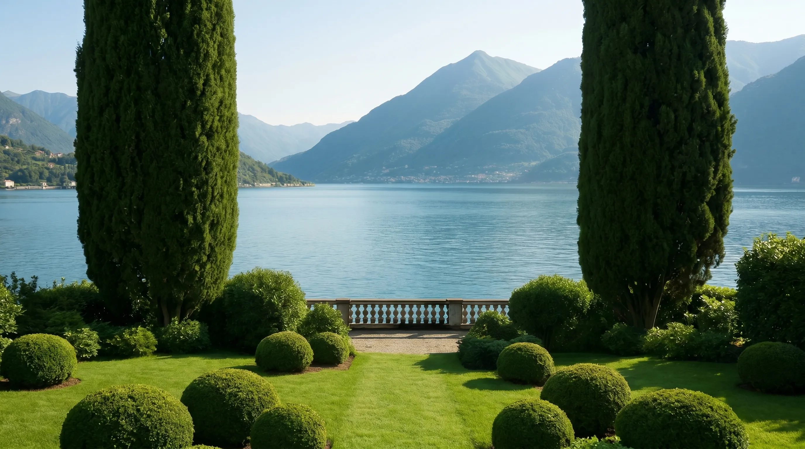 Manicured garden with round shrubs and tall trees overlooking a calm lake and distant mountains.