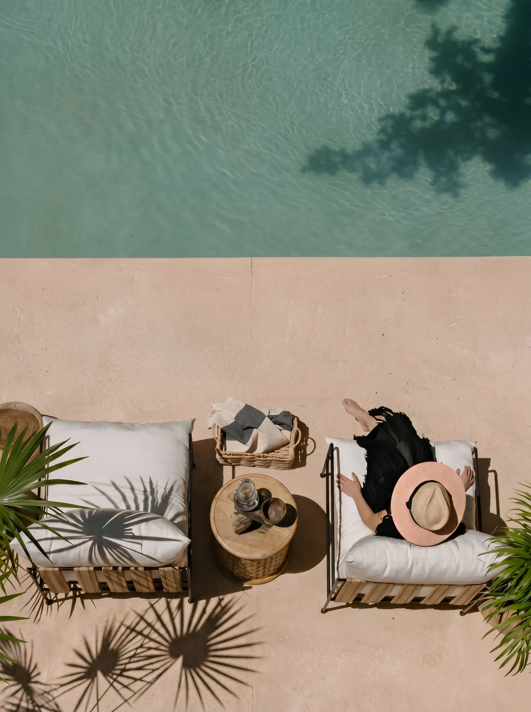 Person in a sun hat lounging on a cushioned chair by a pool with a wooden table and basket nearby.