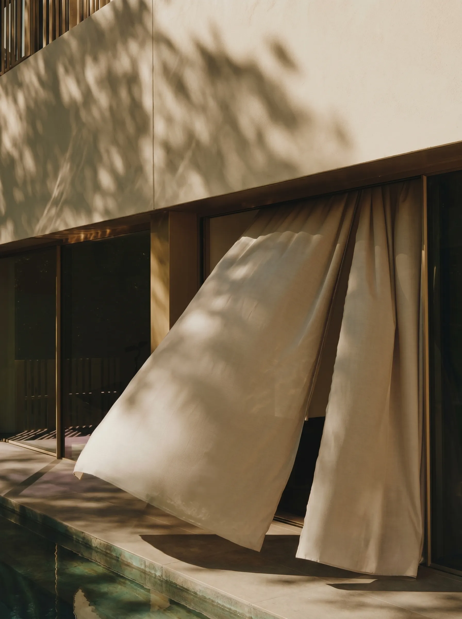 Light beige curtains blowing outward from large glass doors beside a reflecting pool outdoors.