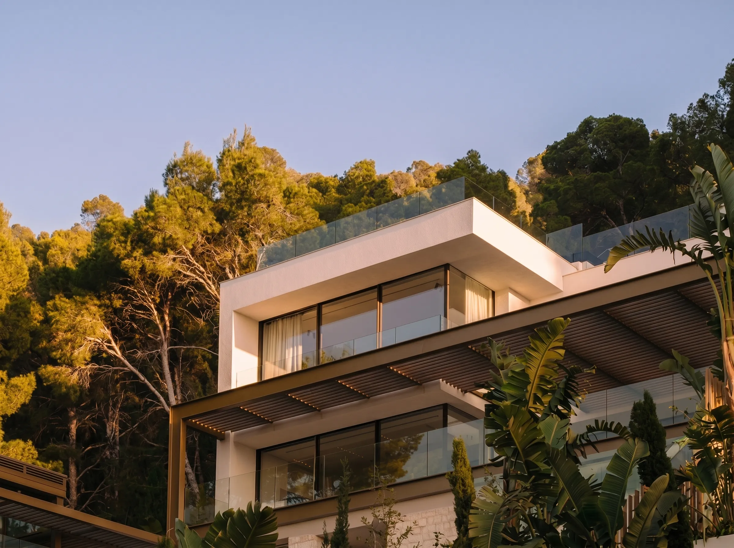 Modern white house with large glass windows and balcony surrounded by trees in soft sunlight.