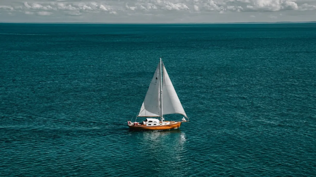 Single sailboat with white sails floating on calm ocean waters under a cloudy sky.