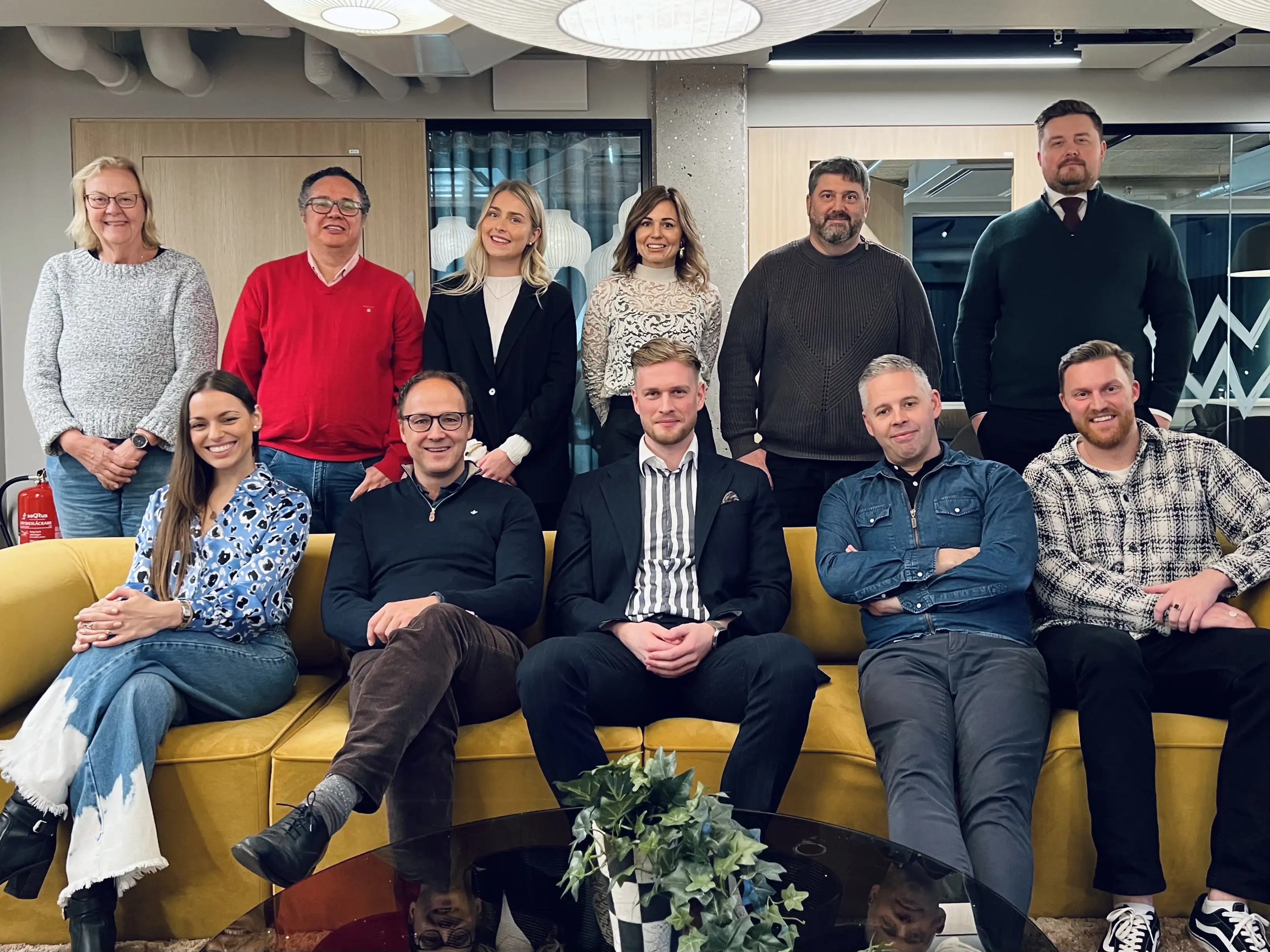 Group of eleven adults posing and smiling in a modern office on a yellow couch and behind it.