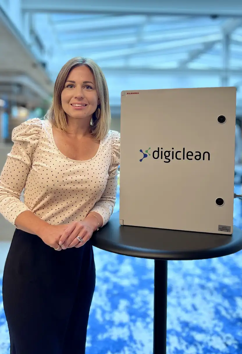 Smiling woman in polka-dot blouse and black skirt standing next to a device labeled digiclean on a table.