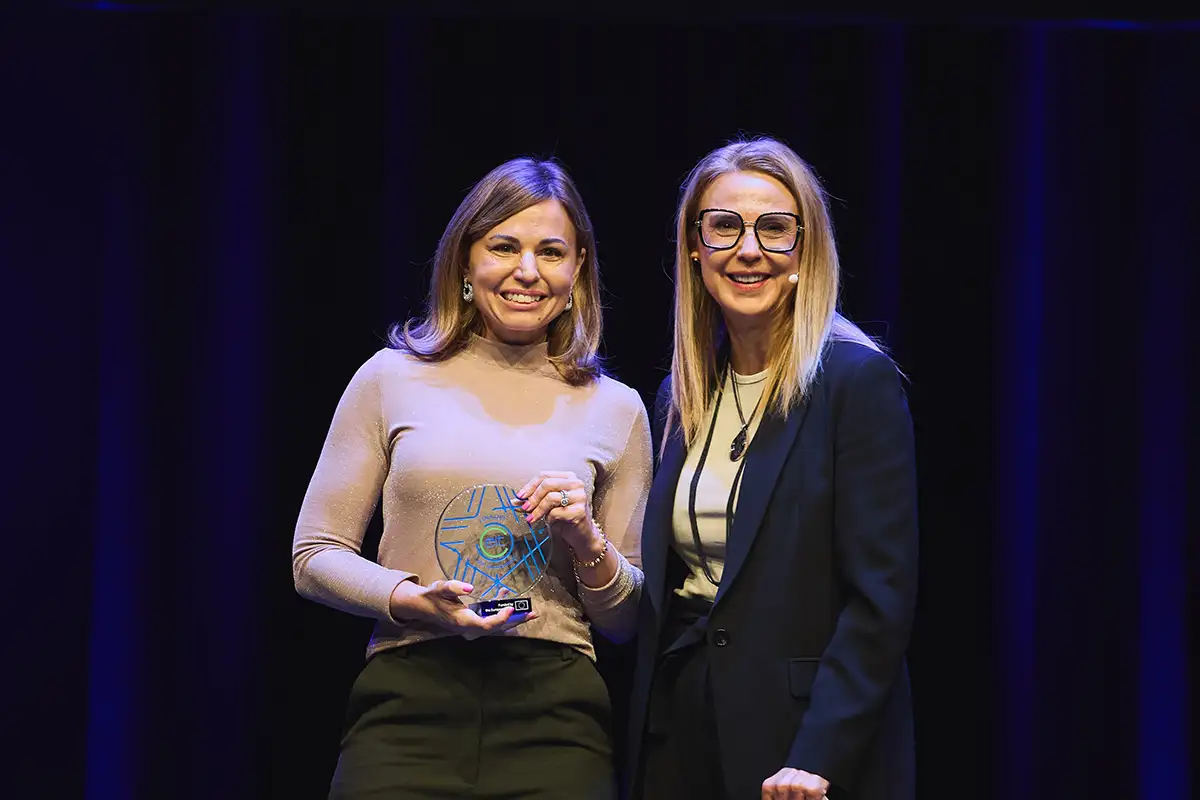 Two women smile on stage; one holds a round glass award with blue designs.