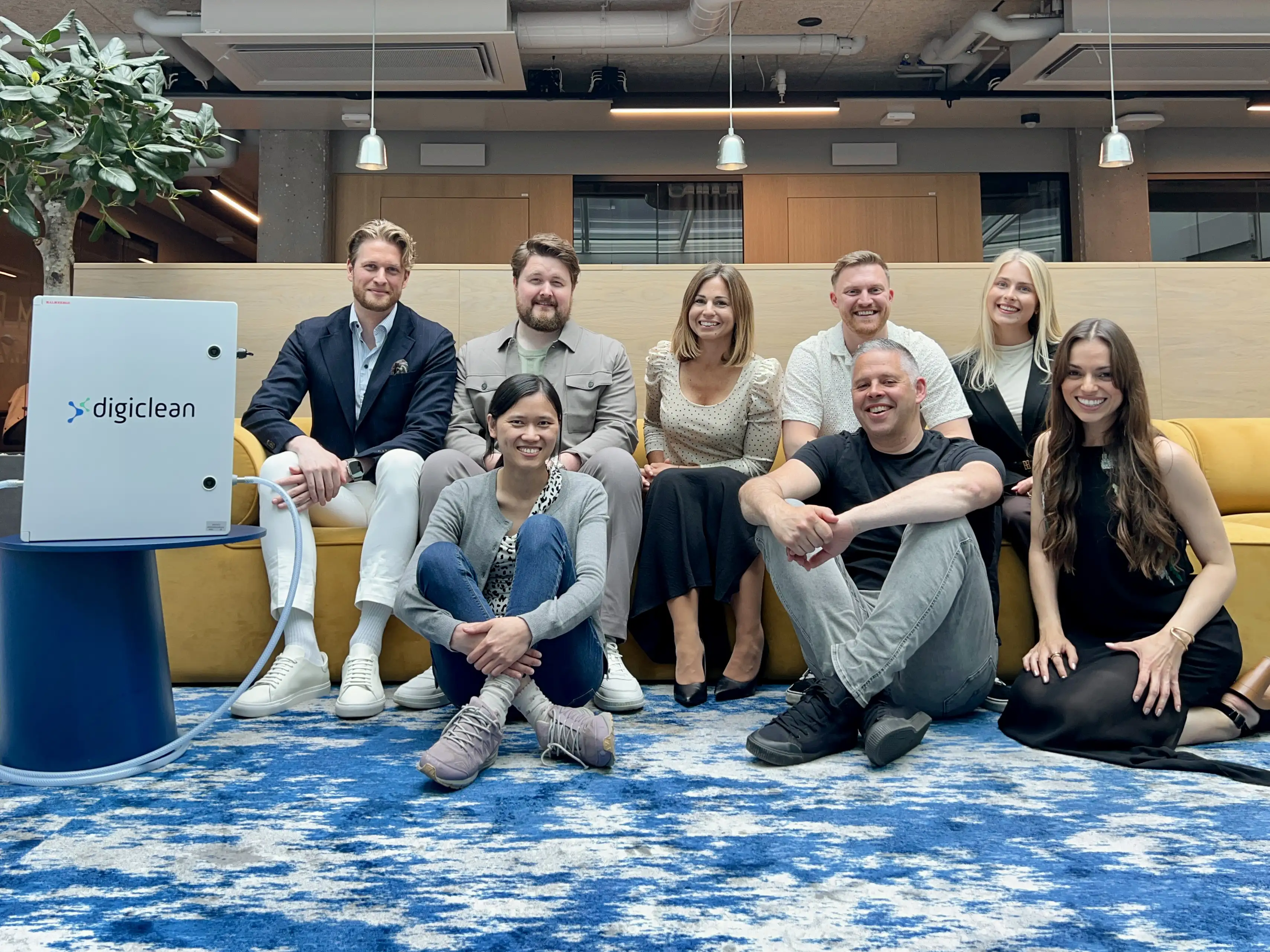 Eight people sitting and kneeling indoors near a yellow couch with a digiclean device on a blue table.