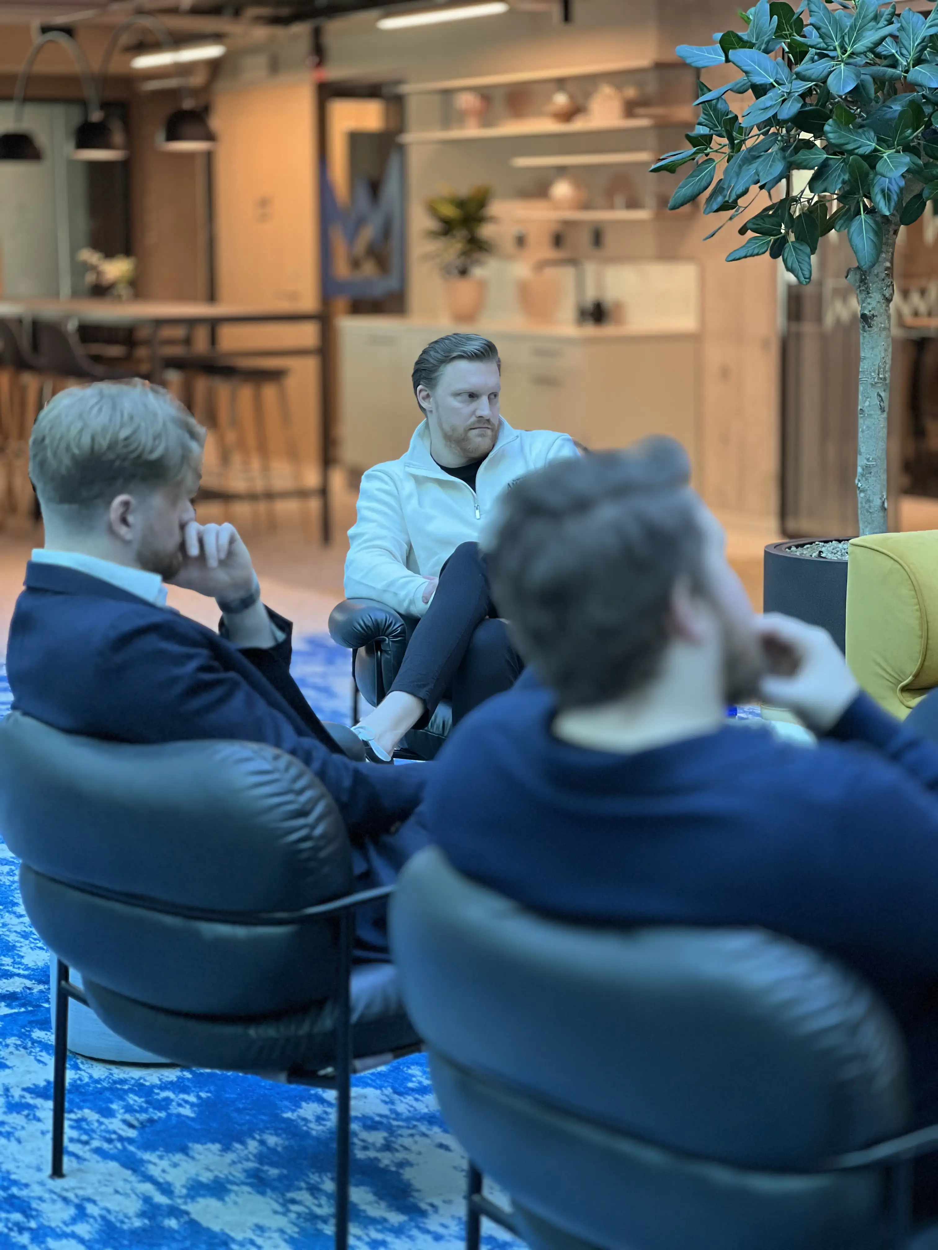 Three men seated in a modern indoor space with blue patterned carpet and a large potted plant.