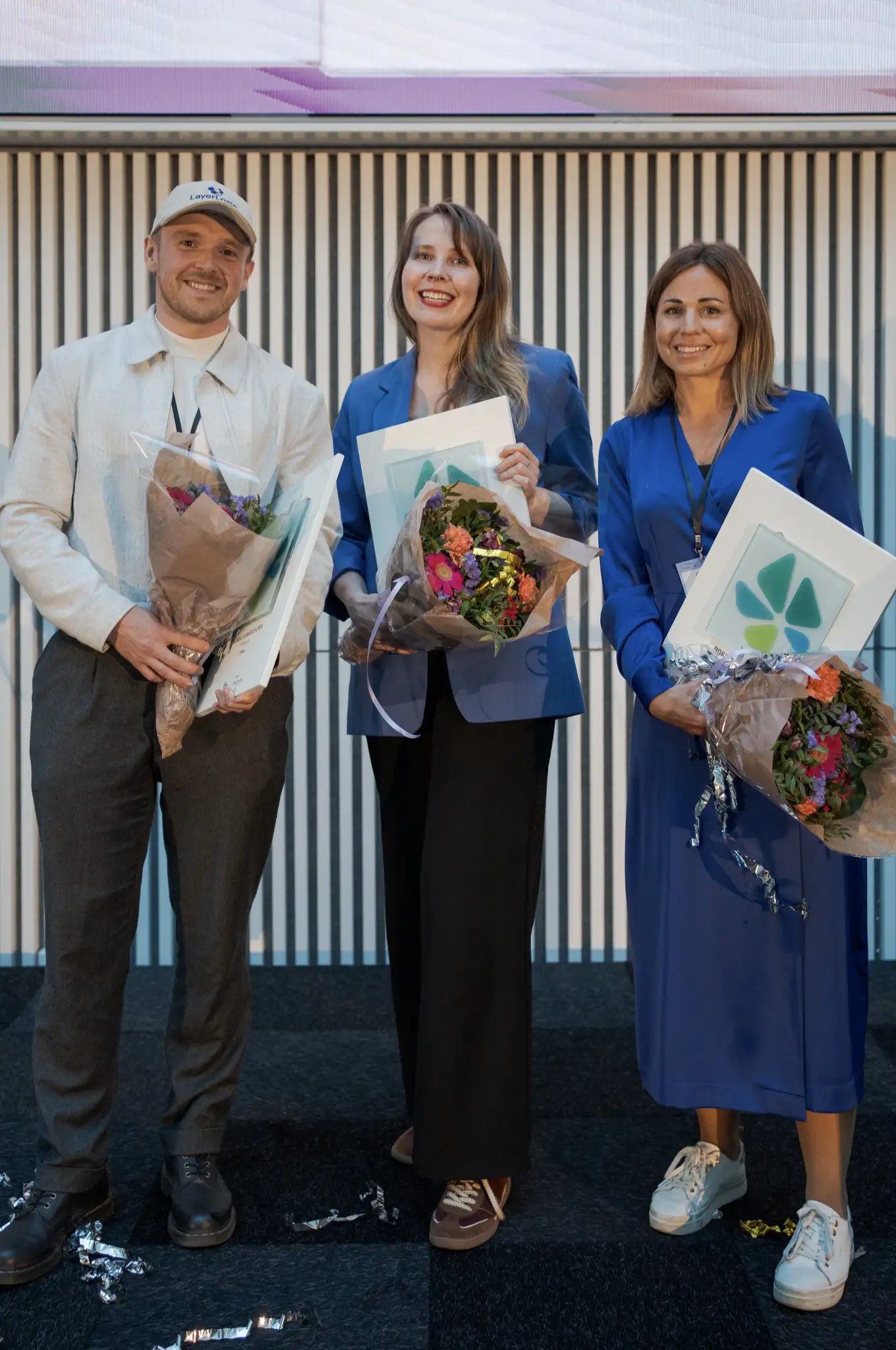 Three people smiling, holding bouquets and framed certificates at an event with a striped background.