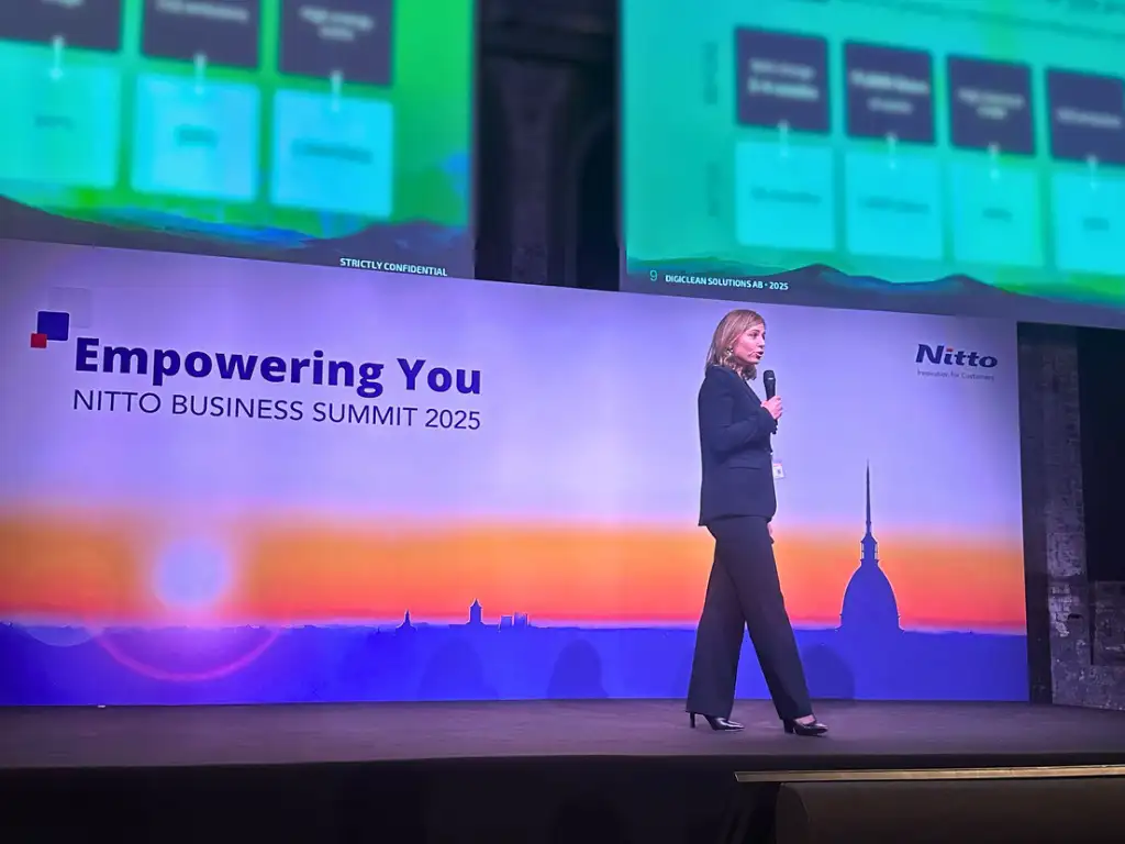 Woman in dark suit speaking on stage at Nitto Business Summit 2025 with Empowering You backdrop.