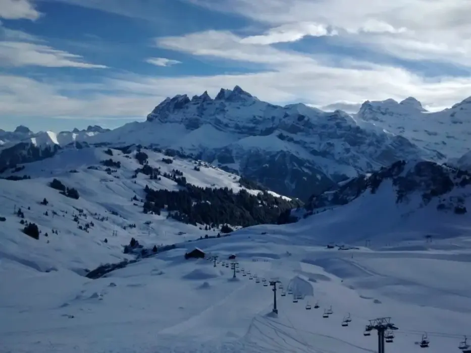 Paysage de montagnes enneigées avec une remontée mécanique et un ciel partiellement nuageux en arrière-plan.