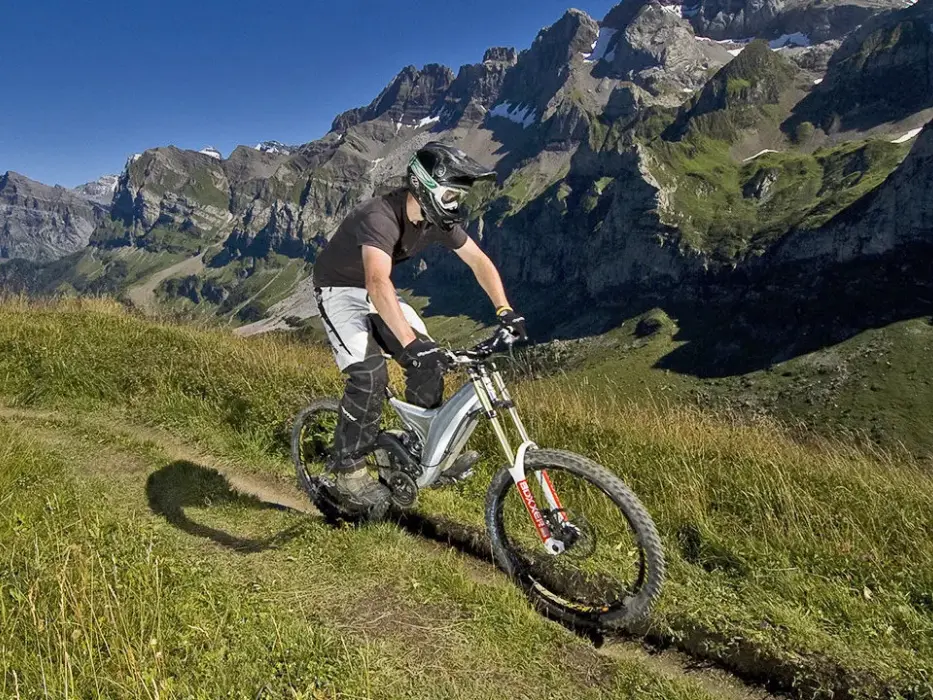 Personne en tenue de protection fait du VTT sur un sentier de montagne herbeux sous un ciel bleu.