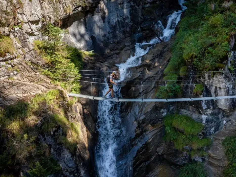Un randonneur traverse un pont suspendu au-dessus d'une cascade rocheuse en montagne.