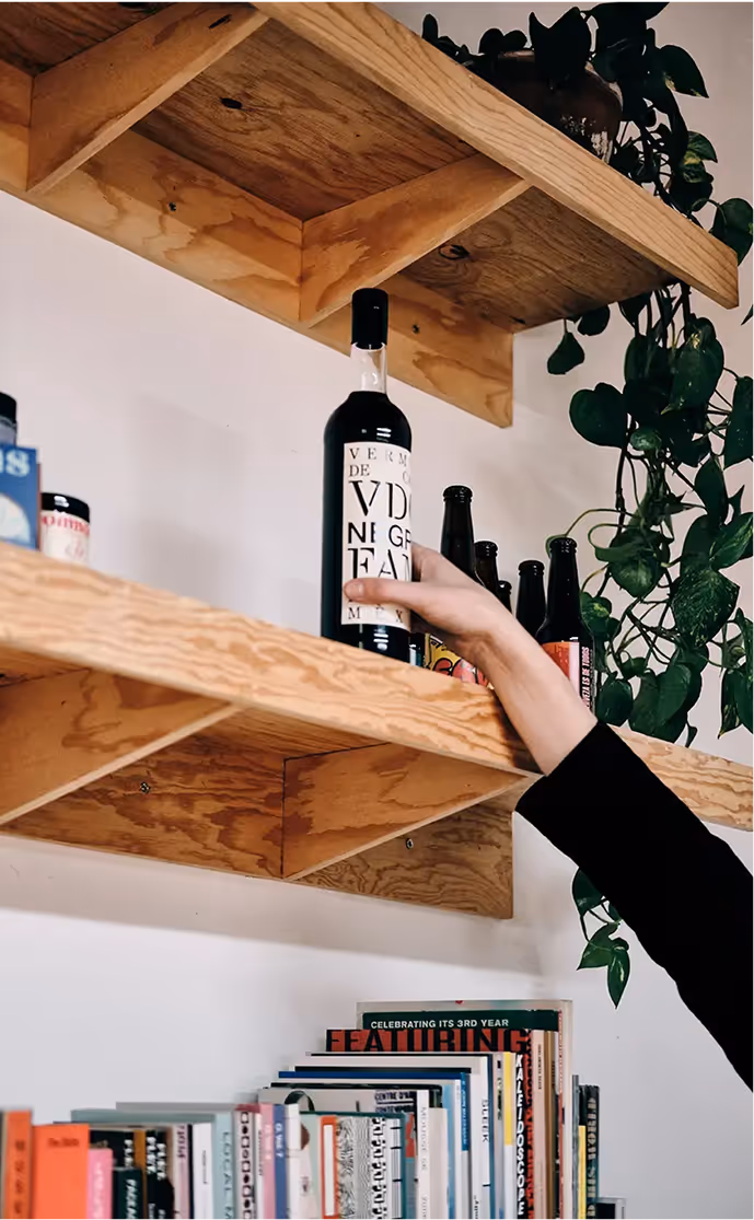 Hand reaching to grab a tall bottle with a white label from a wooden shelf with more bottles and a green plant hanging nearby.