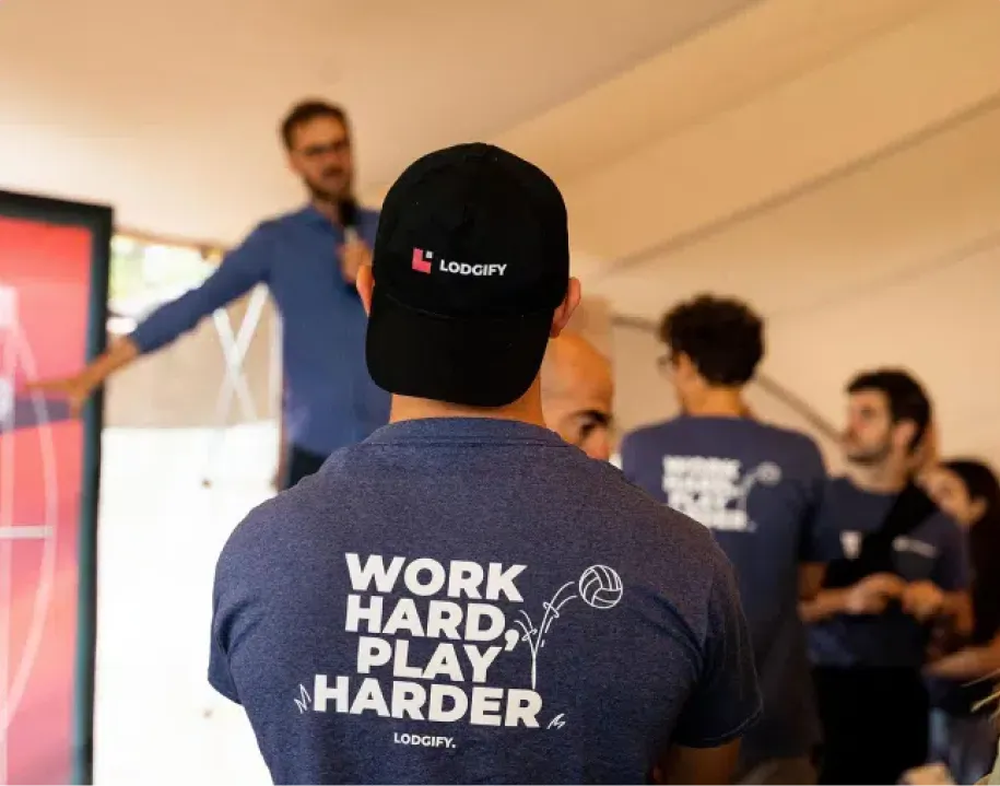 Person wearing a black cap with Lodgify logo and a navy shirt with 'WORK HARD, PLAY HARDER' text and volleyball graphic, in a group setting.