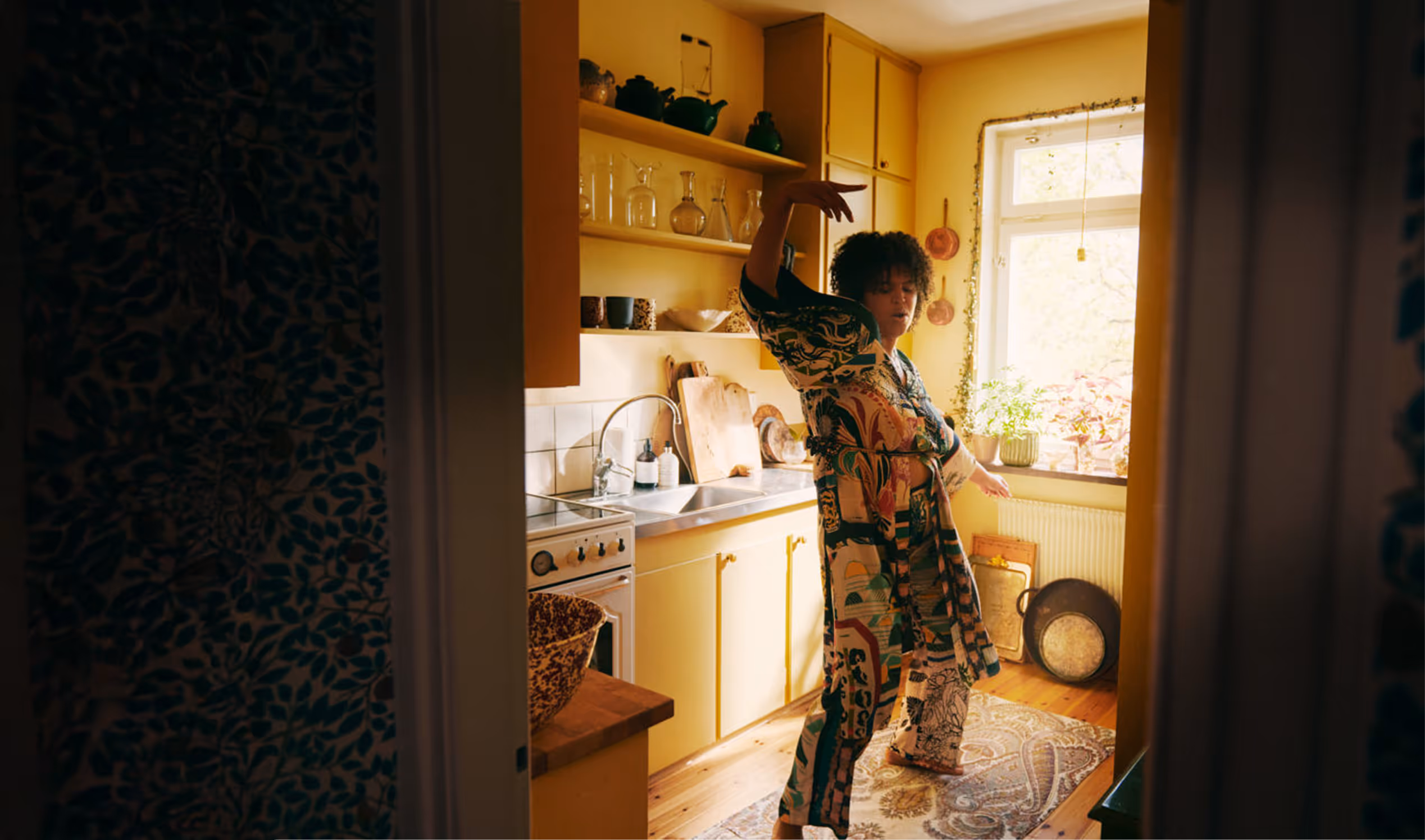 Person in colorful patterned clothing dancing in a cozy yellow kitchen with wooden floor and plants on the windowsill.