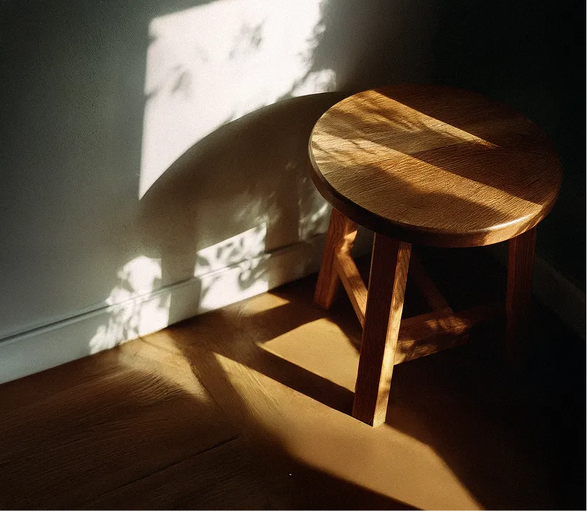Wooden stool casting shadows on a wooden floor with dappled sunlight and plant shadows on the wall.