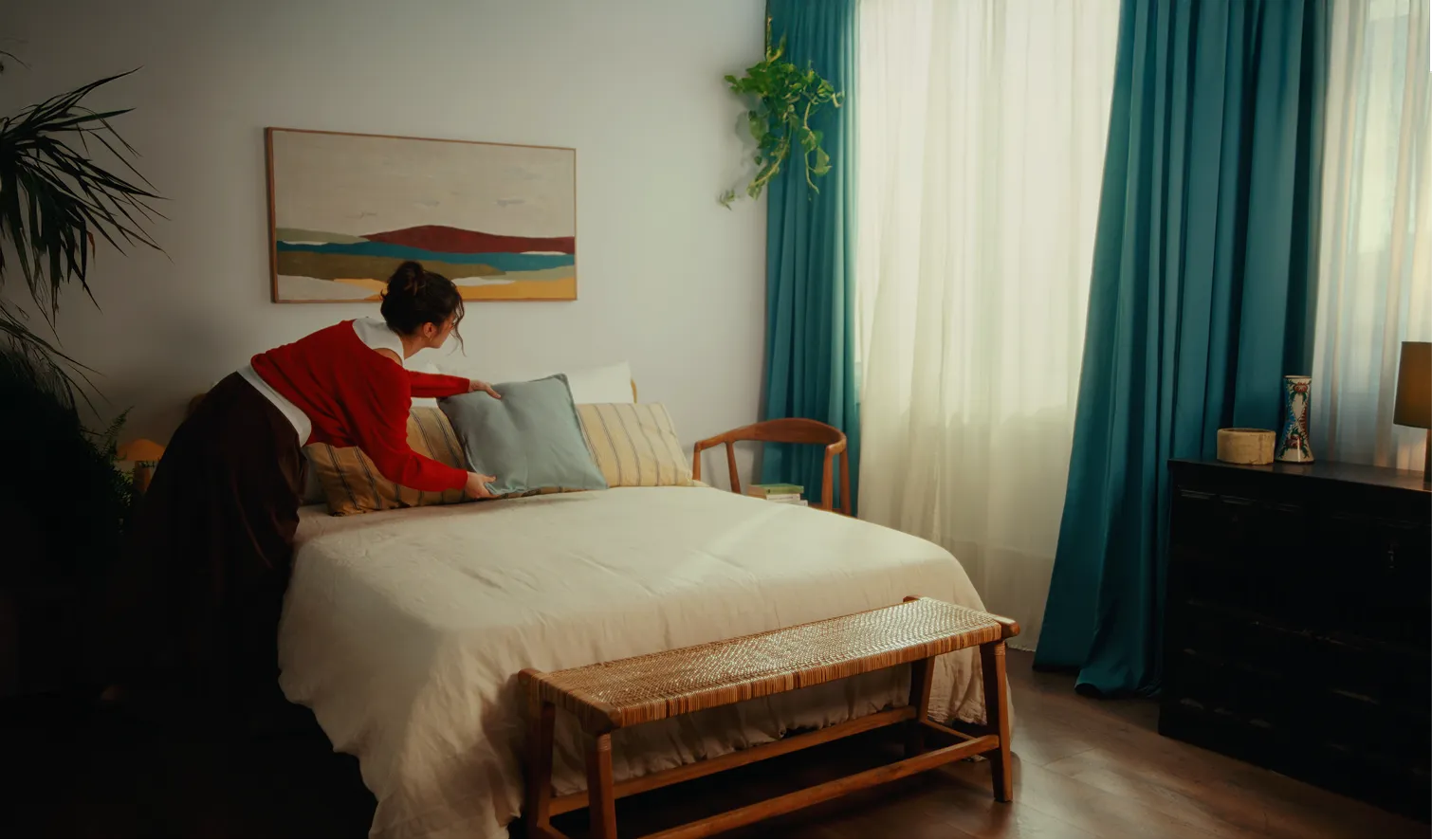 Woman arranging a light blue pillow on a neatly made bed with beige bedding in a bedroom with blue curtains and wooden furniture.