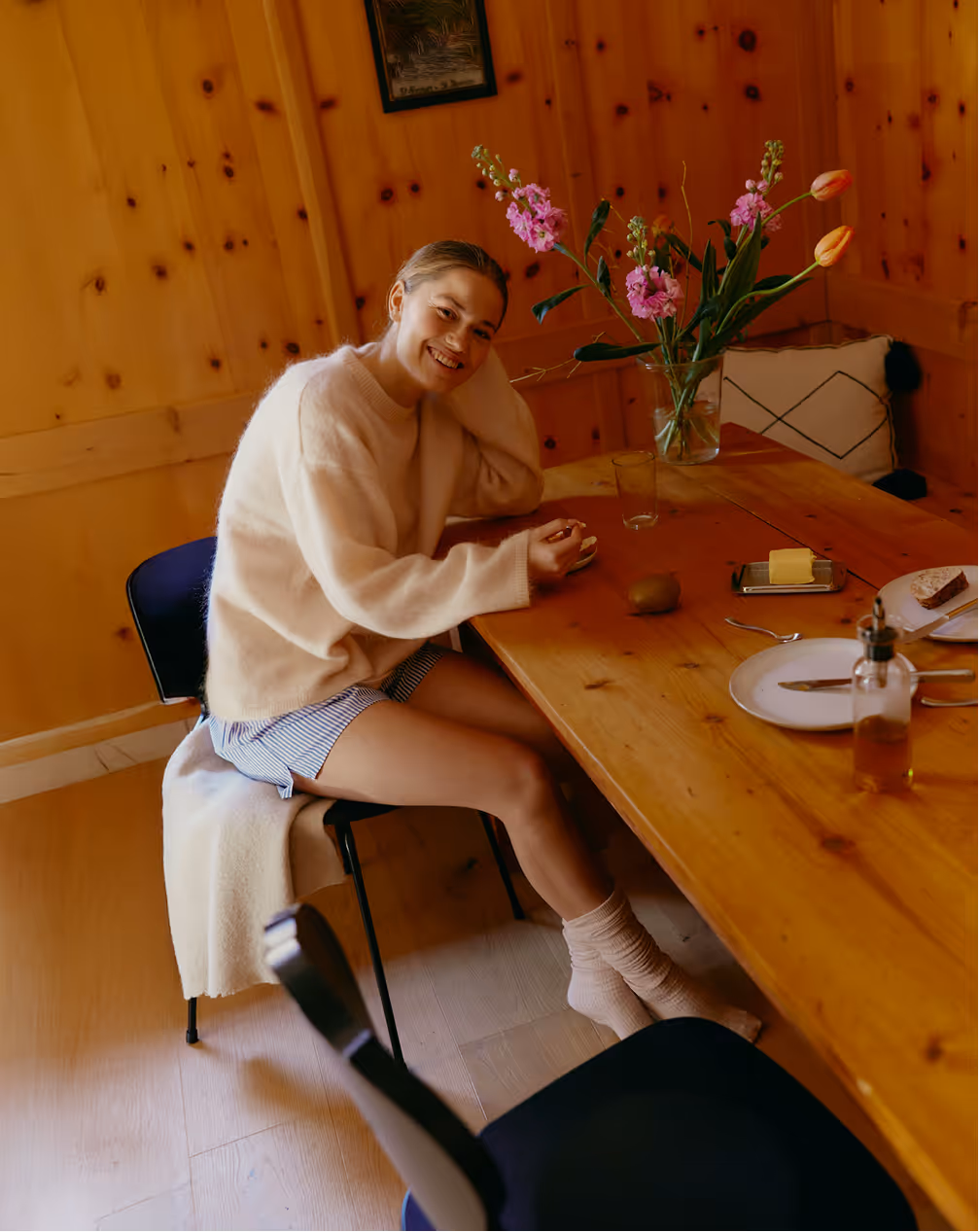 Smiling woman in cozy sweater and socks sitting at wooden table with flowers, plates, and food in a warmly lit room.