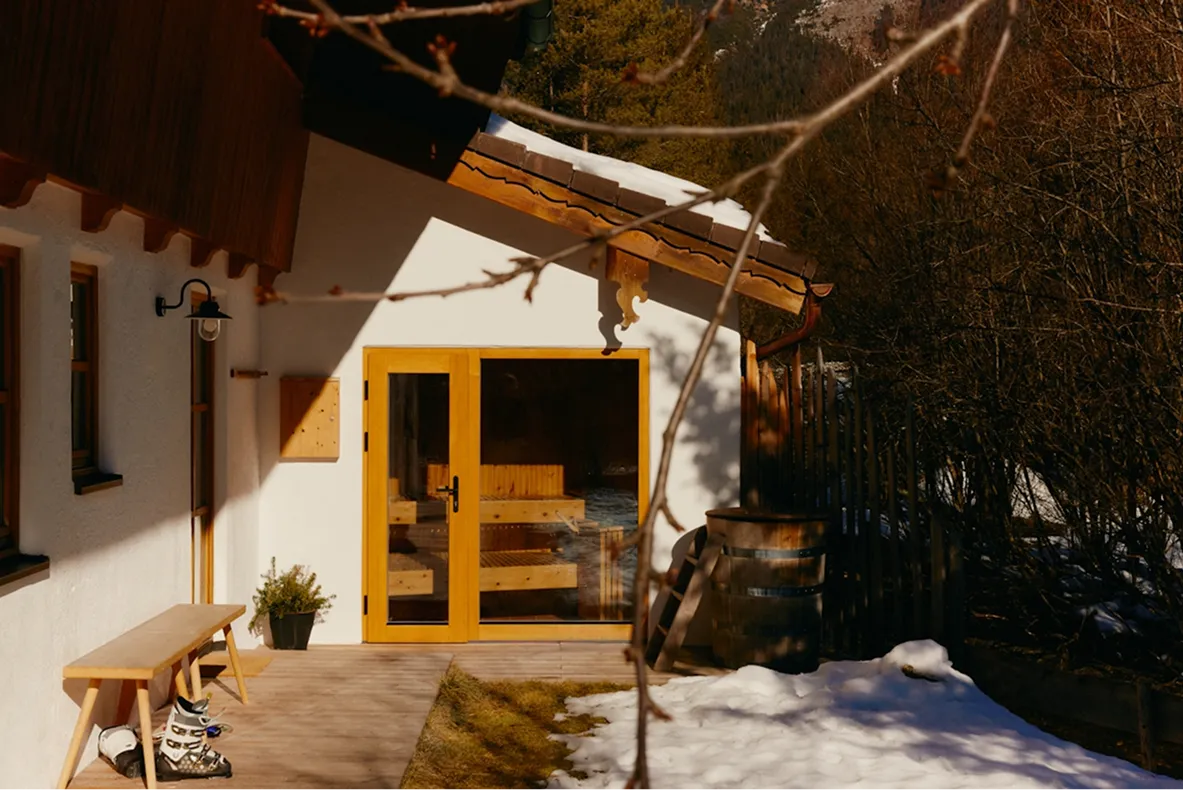 Sauna cabin with wooden bench, ski boots, and partially snow-covered ground outside in a mountain setting.