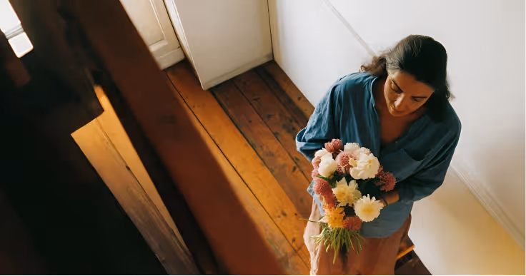 Woman in a blue shirt holding a bouquet of mixed flowers, standing on wooden floor near white walls.