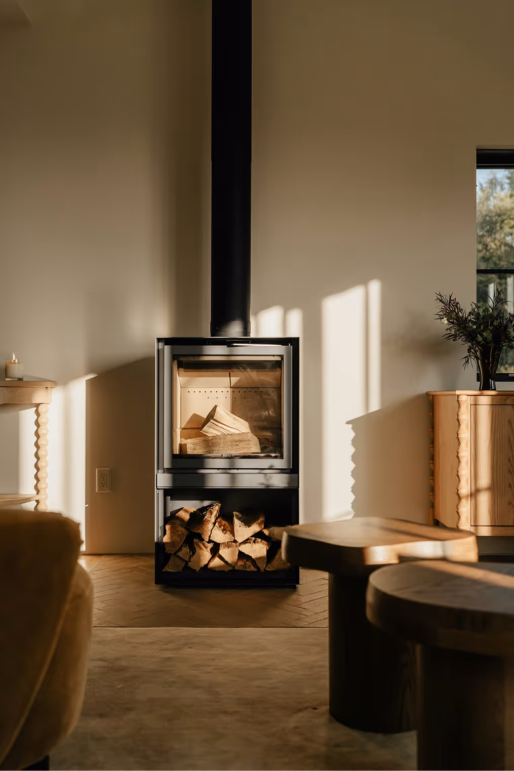 Modern black wood-burning stove with firewood inside and stored below in a warmly lit, minimal living room.