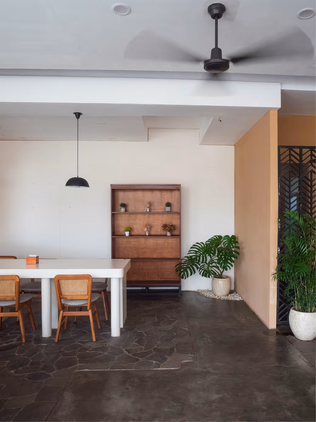 Modern dining area with a white table, four wooden chairs, a wooden shelf with small plants, and two large potted plants on a stone and concrete floor.