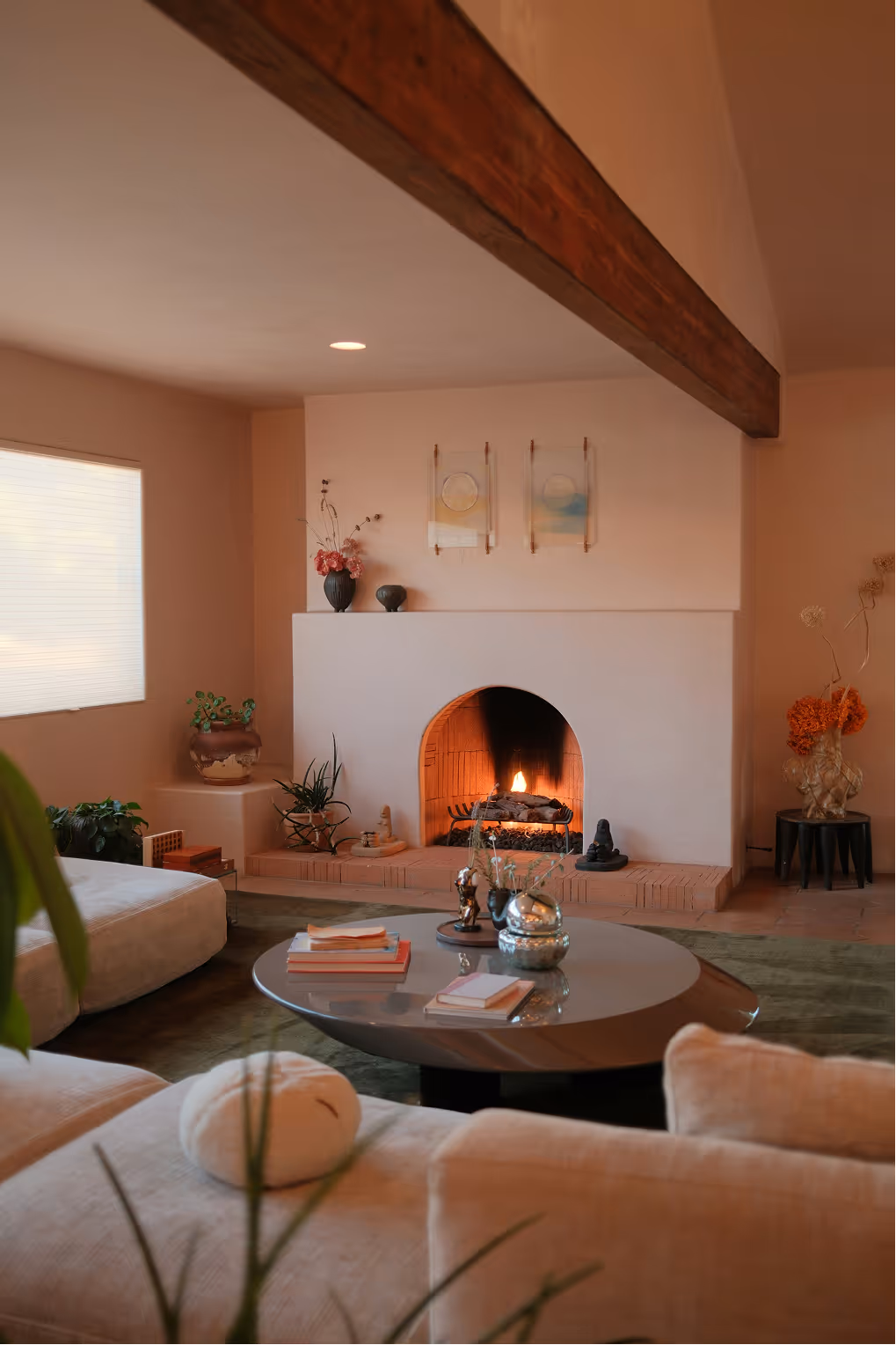 Cozy living room with modern furniture, a round coffee table with books, and a lit fireplace with plants and decorative items around.
