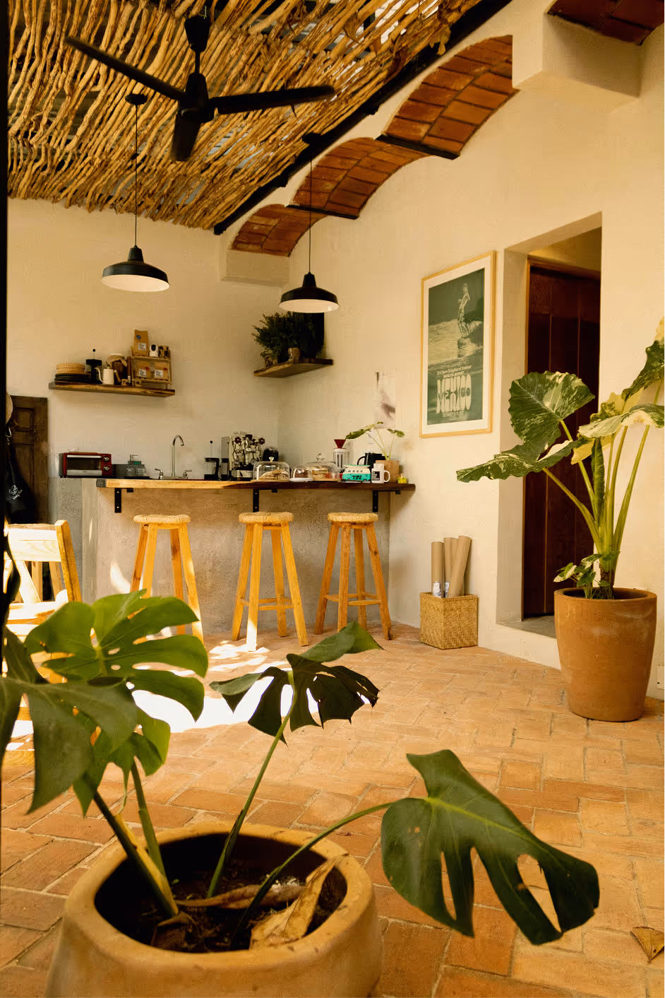 Cozy indoor space with wooden stools at a bar counter, potted plants, hanging lights, and a rustic ceiling made of intertwined branches.