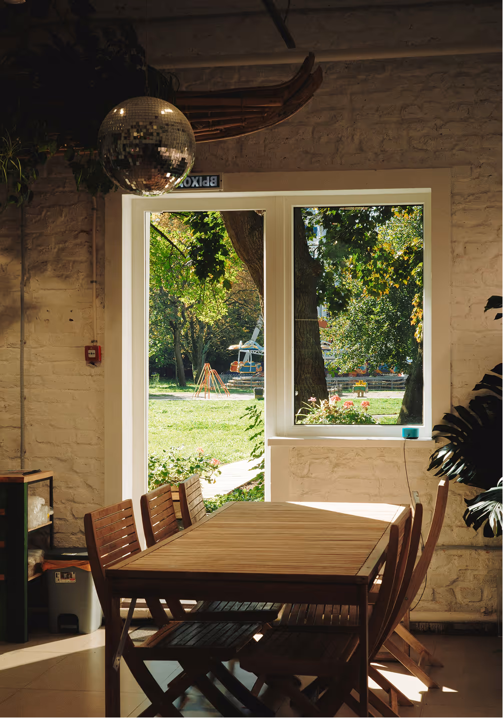 Sunlit wooden dining table with chairs inside a room facing a window and open door showing green trees and playground outside.