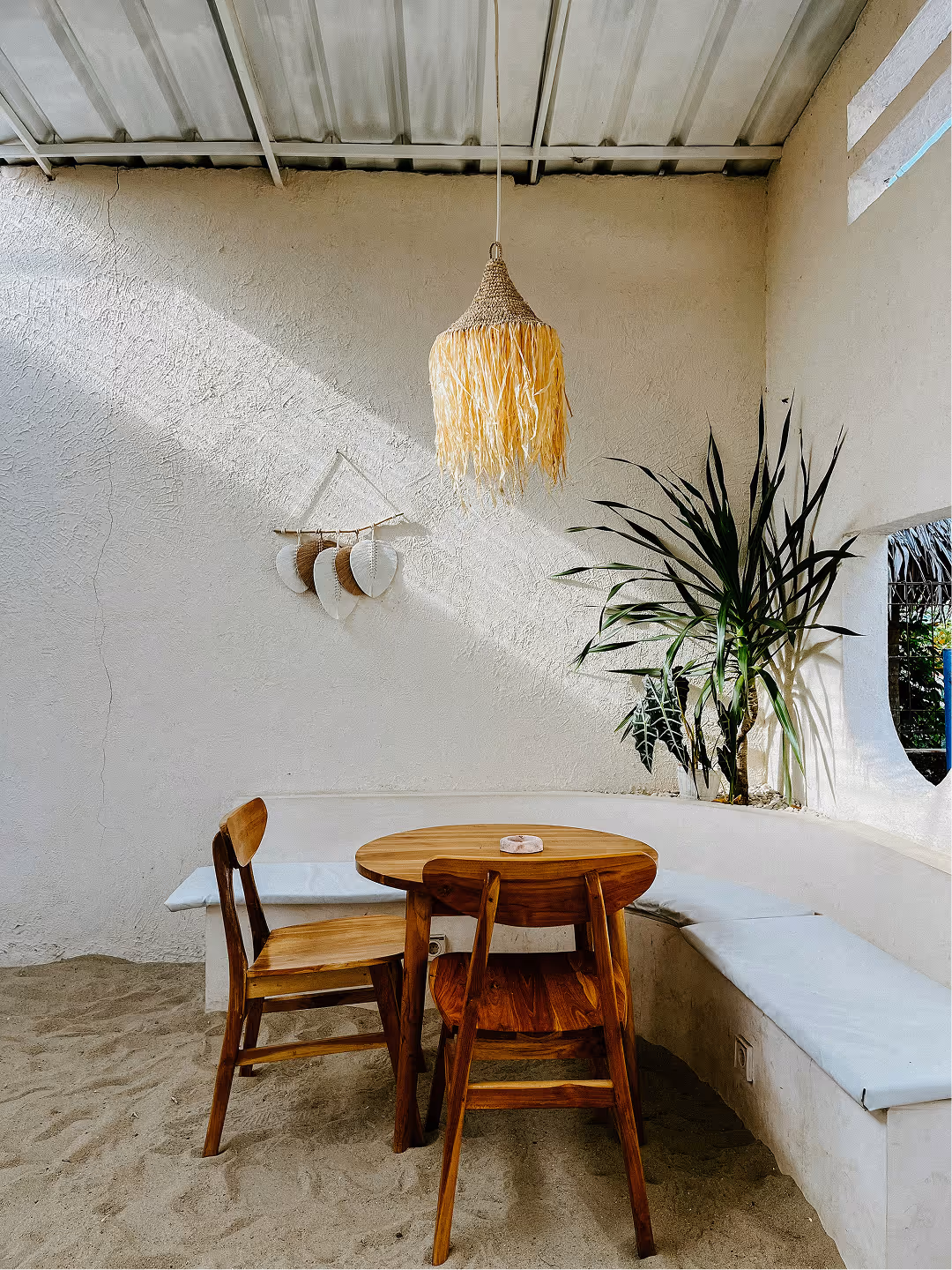 Cozy seating area with wooden table and chairs on sandy floor, white cushioned bench, hanging woven light fixture, and potted plants under a metal roof.