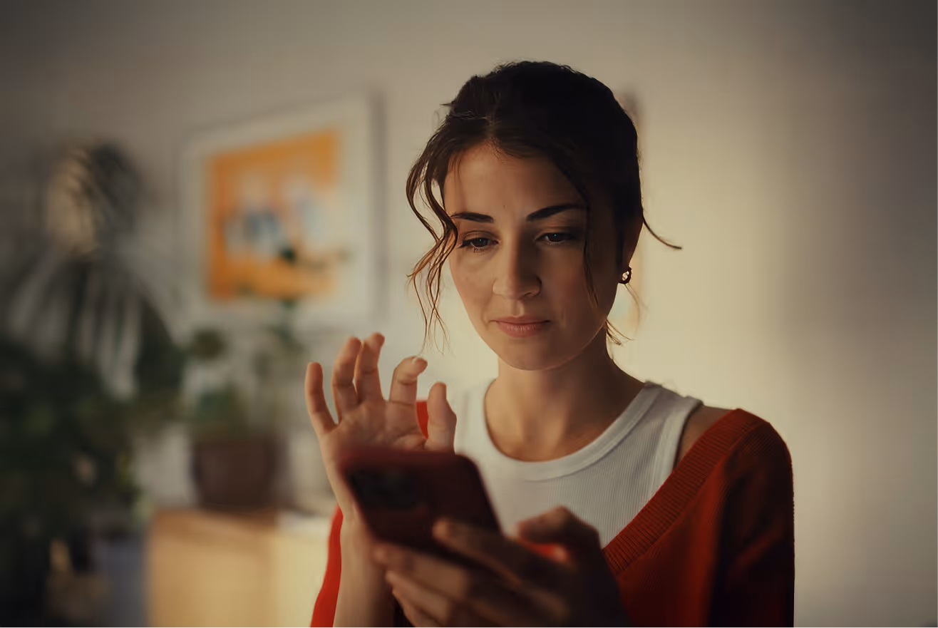 Young woman with dark hair wearing red and white, looking intently at a smartphone in a softly lit room.