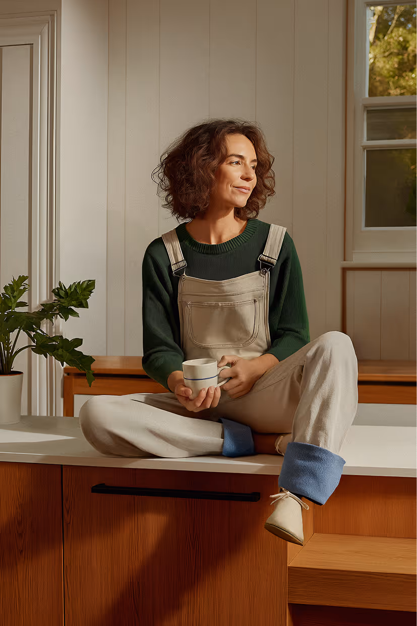 Young woman with curly hair sitting cross-legged on a kitchen counter, holding a mug and looking to the side, wearing beige overalls and a green sweater.