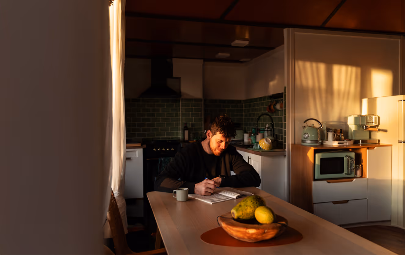 Man writing in a notebook at a kitchen table with a cup and a bowl of fruit, illuminated by warm sunlight.